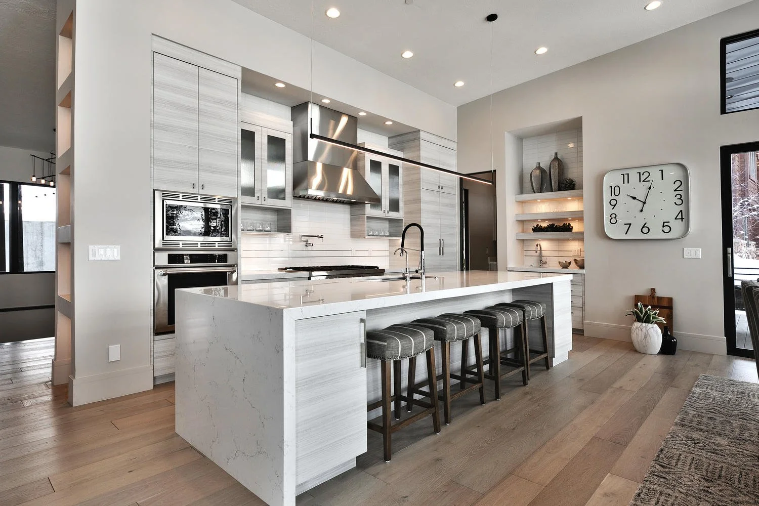 Modern kitchen with white marble island, stainless steel appliances, gray wooden cabinets, and a large wall clock.