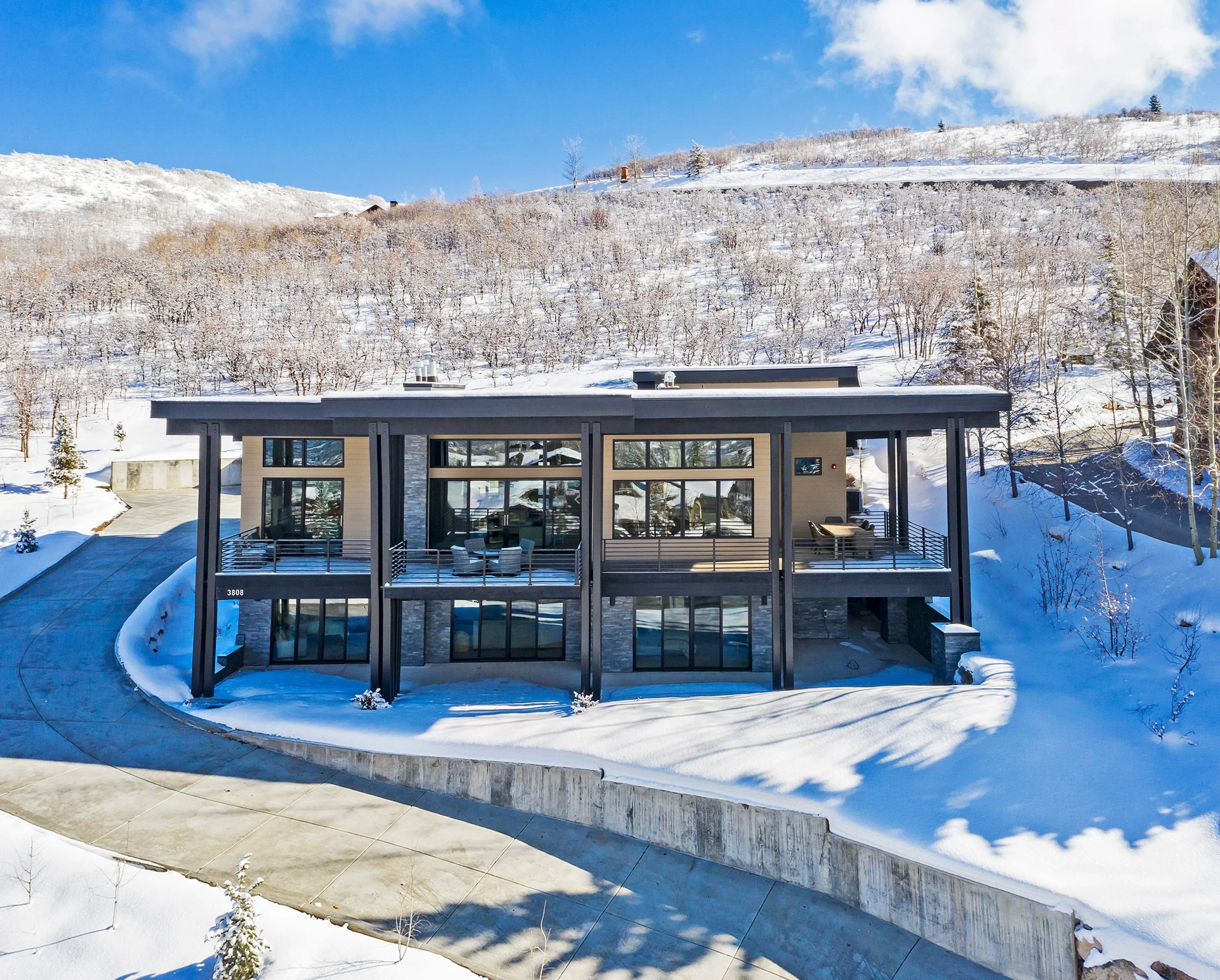 Modern house with large glass windows and a flat roof, surrounded by snow-covered ground and trees, with a snow-capped hill and blue sky in the background.