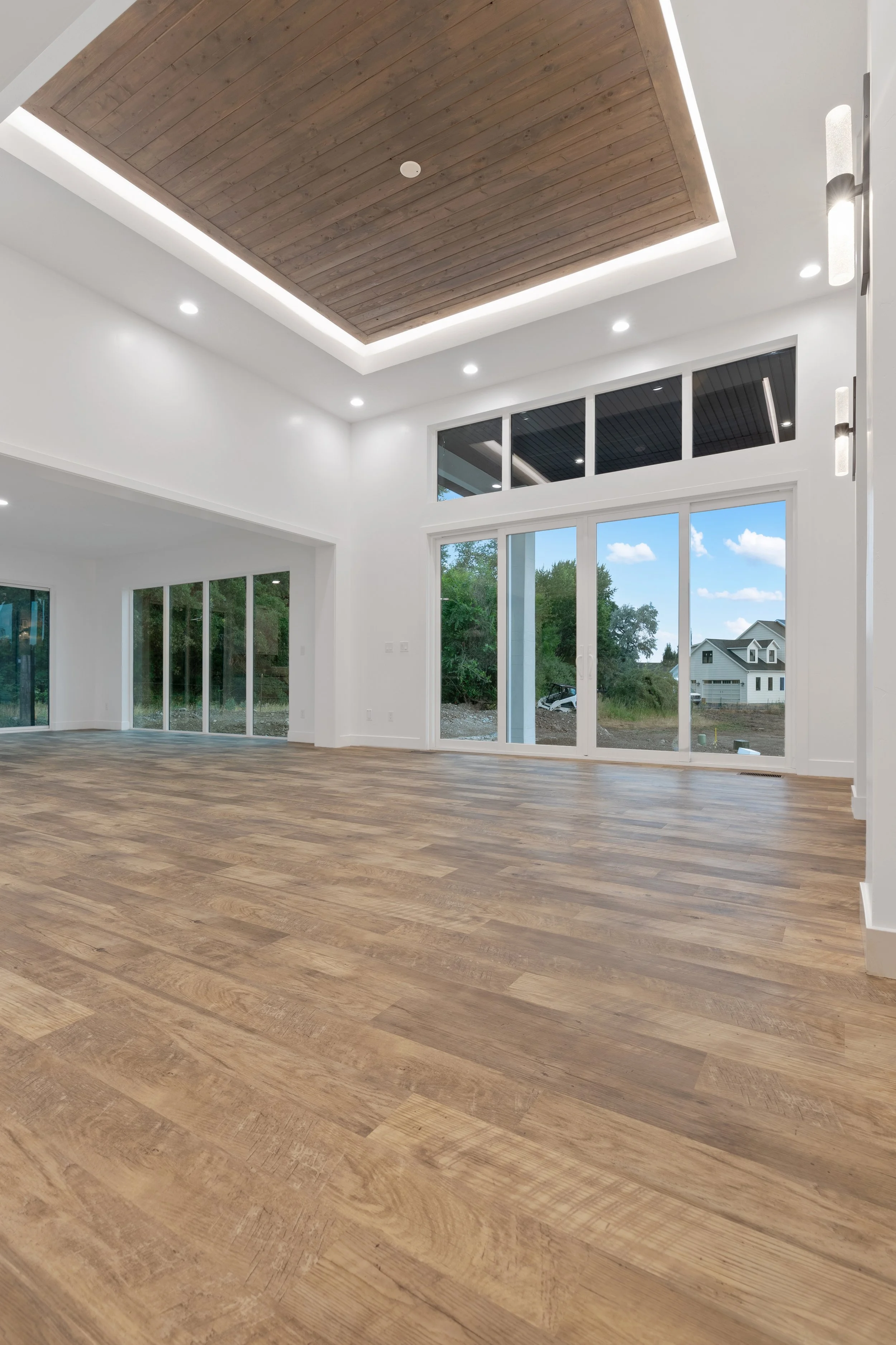Empty modern living room with large windows, wooden flooring, white walls, and a ceiling featuring a wooden inset with recessed lighting.