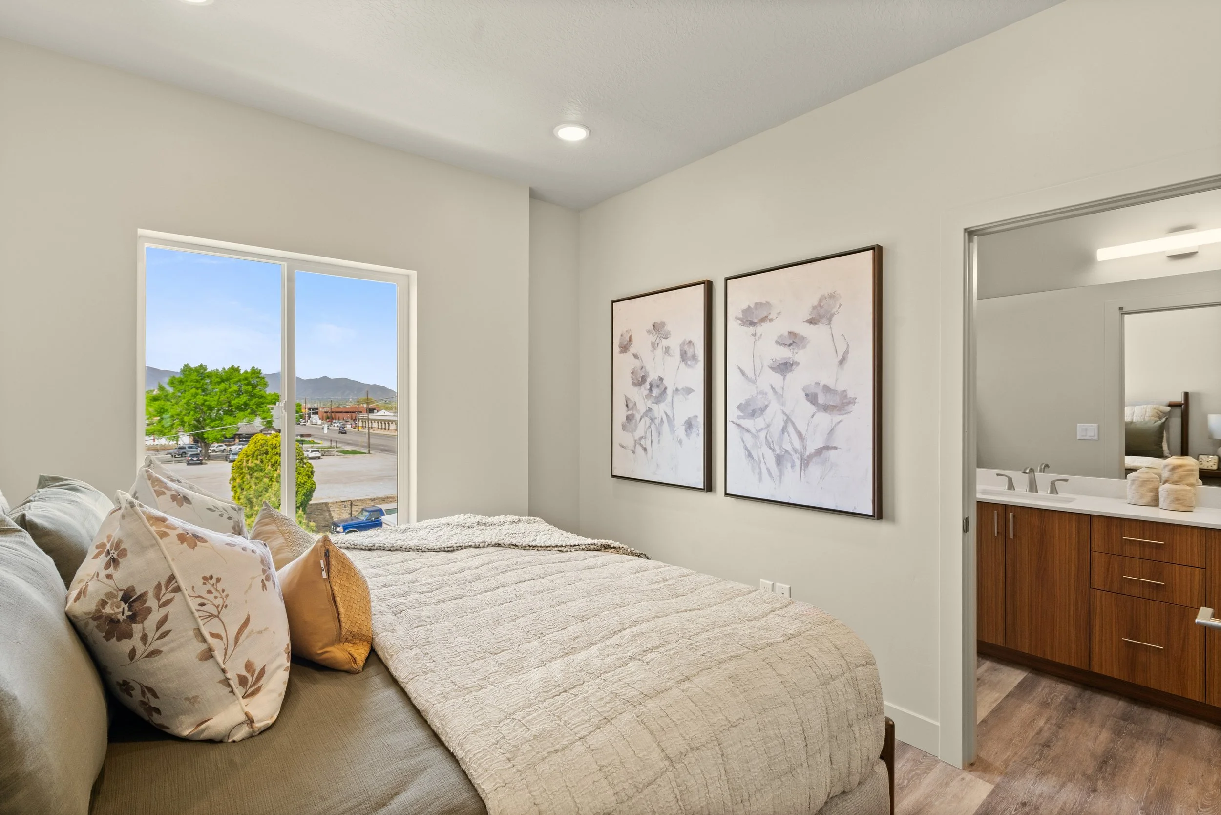 A bedroom with a large bed covered with a beige quilt and decorated with floral pillows. There are two framed botanical prints on the wall, a window showing a tree and distant mountains, and part of a bathroom with wooden cabinetry visible through a doorway.