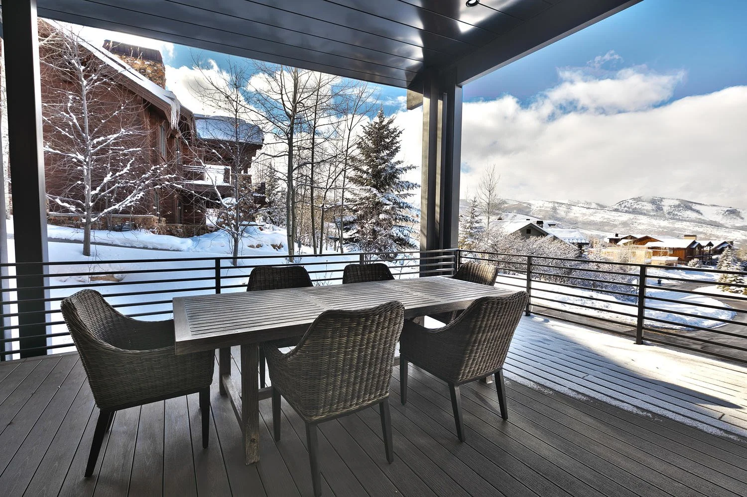 Outdoor balcony with a wooden table and six wicker chairs, overlooking a snowy landscape with houses, trees, and mountains under a partly cloudy sky.
