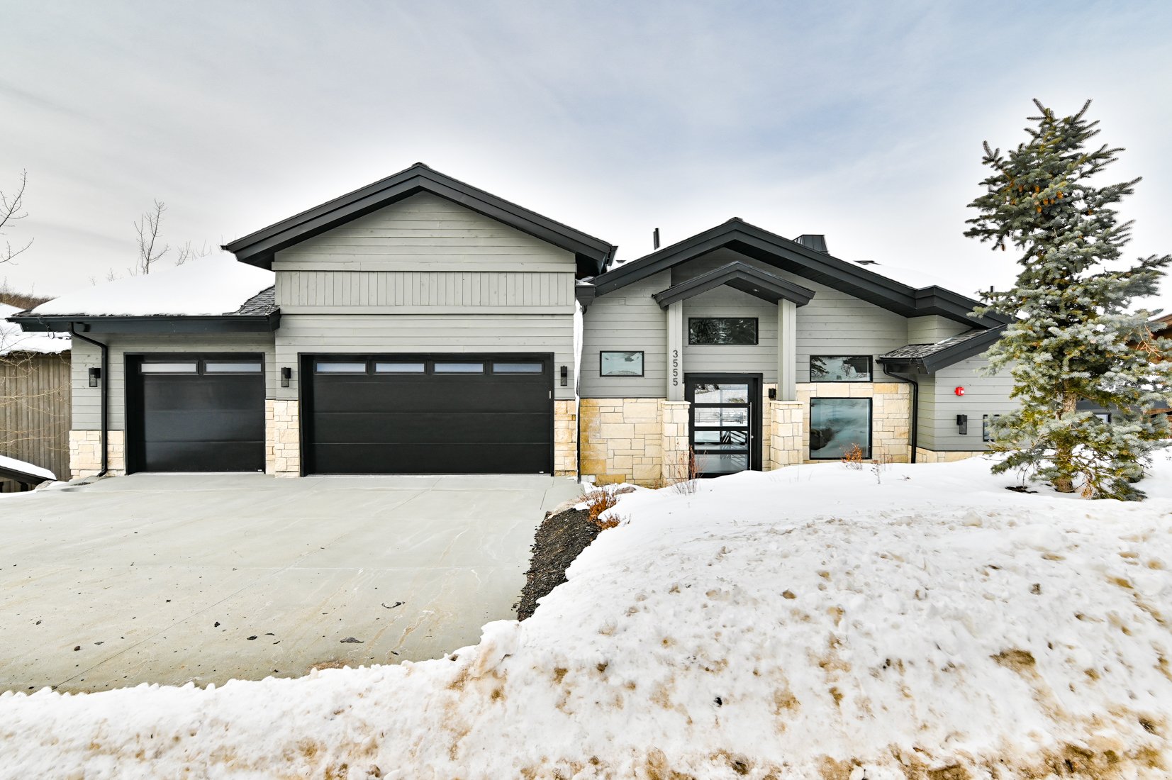 A modern house with black garage doors, stone accents, large windows, and a snow-covered yard with a Christmas tree on the right side.