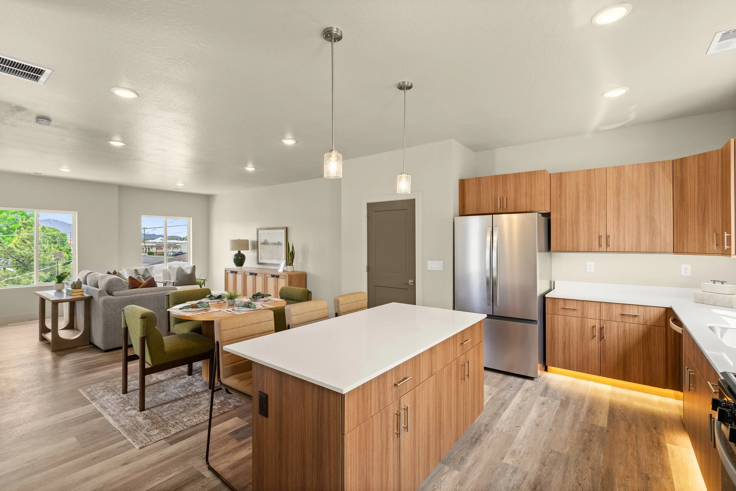 Open-concept kitchen and living room with light wood cabinets, stainless steel refrigerator, white countertops, dining table, and seating area near windows showing outdoor greenery.