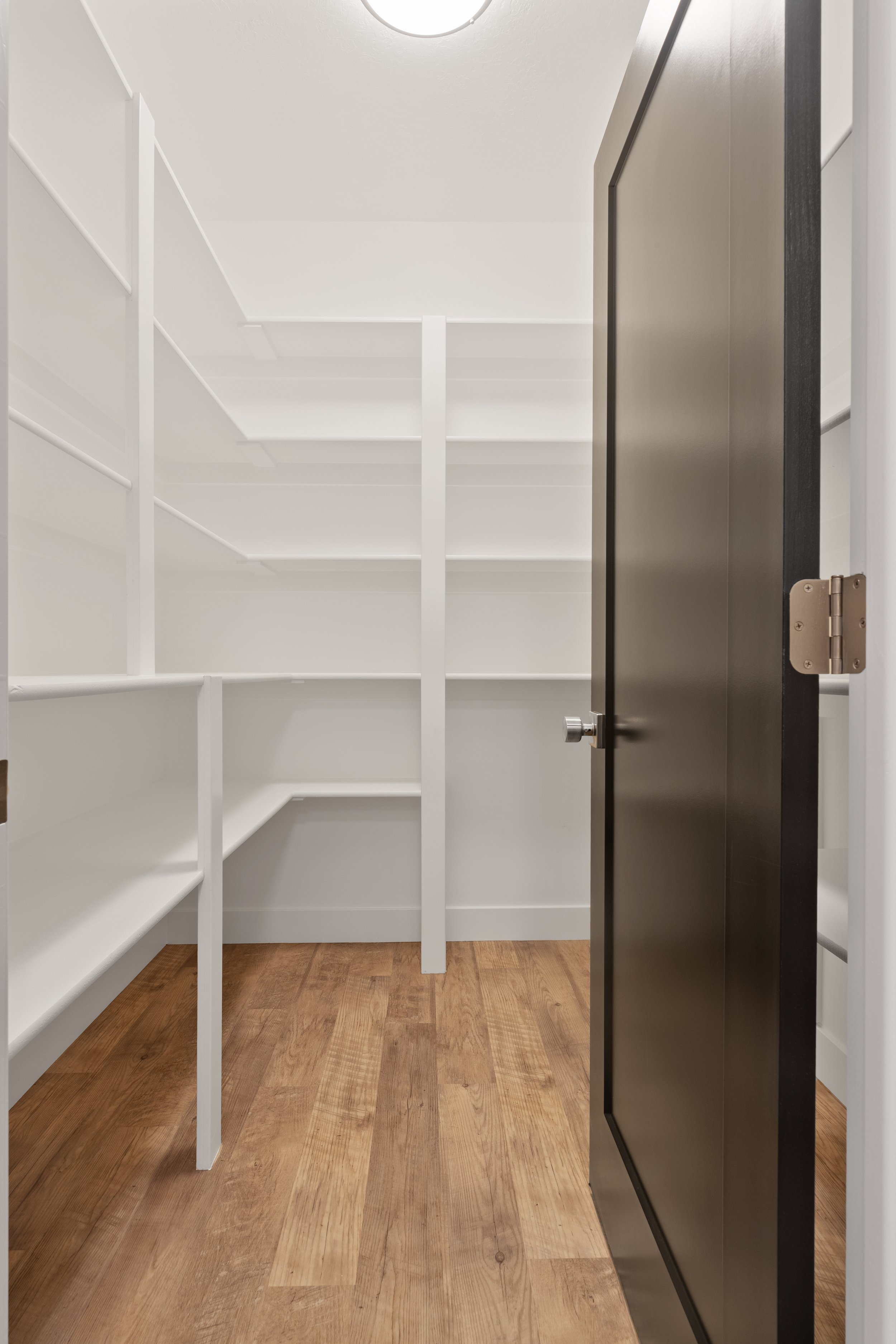 Empty walk-in closet with white shelves on both sides, a closed dark door on the right, hardwood floor, and a ceiling light overhead.