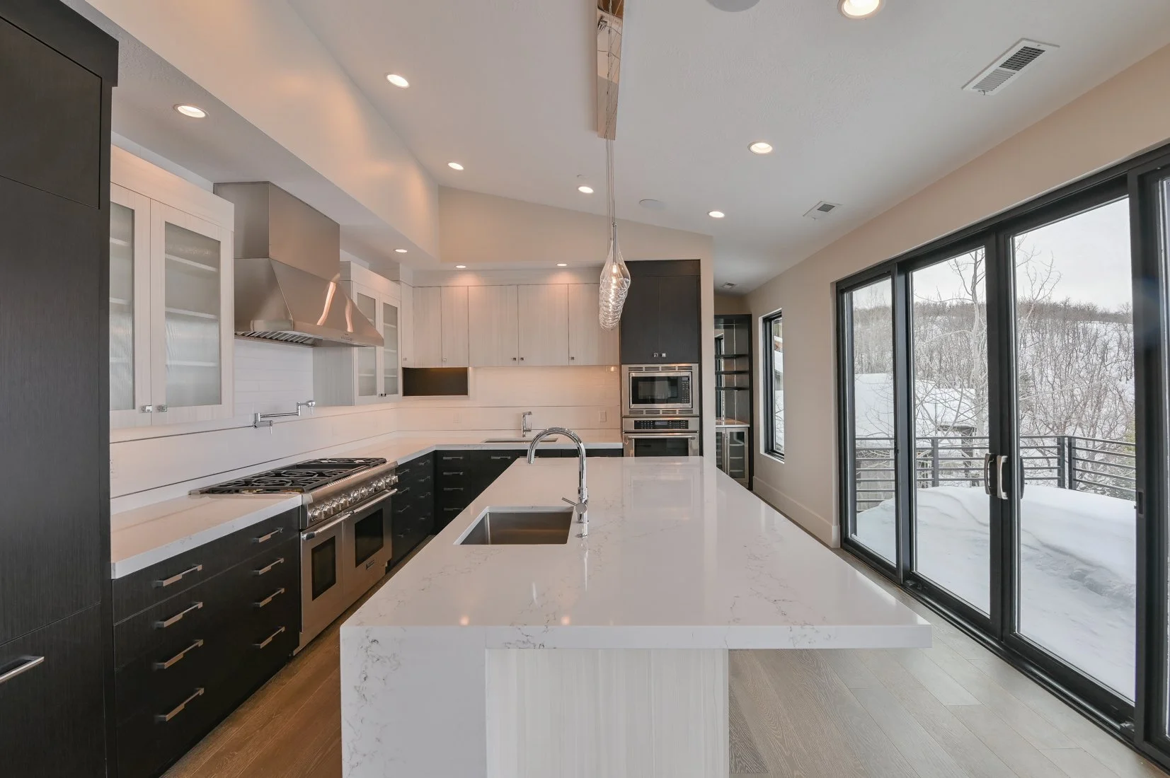 Modern kitchen with white countertops, black lower cabinets, white upper cabinets with glass fronts, stainless steel appliances; large sliding glass doors showing a snowy outdoor scene.
