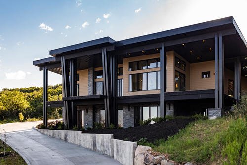 Modern two-story house with large windows and black accents, situated on a hillside with a concrete driveway and green landscaping.