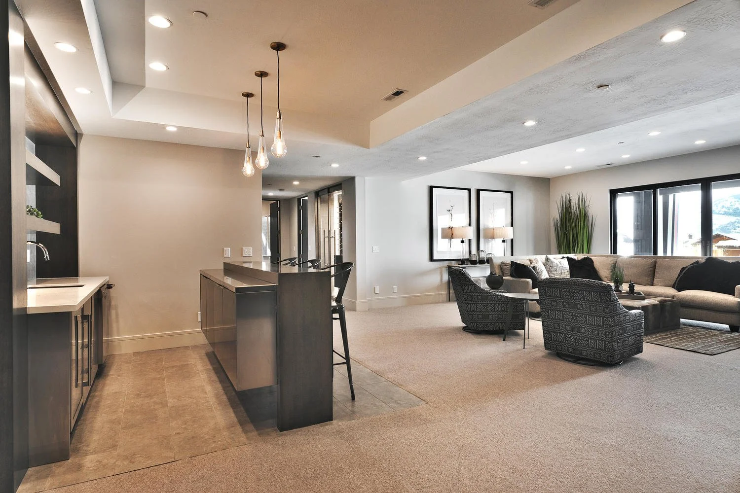 Open living room with large window, beige sofa, black patterned chairs, and coffee table, open kitchen with island and hanging light fixtures, carpeted and tiled flooring.