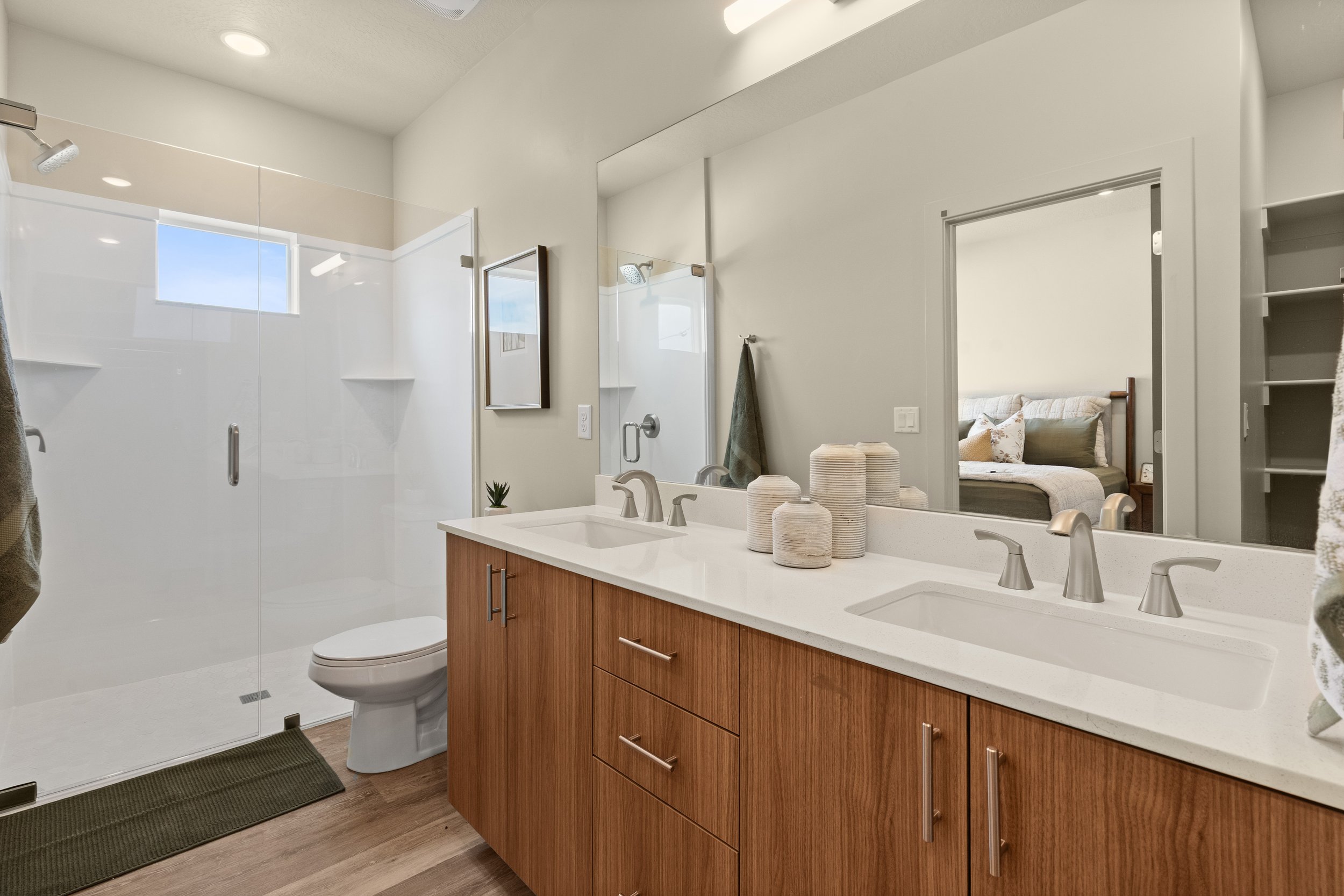 Modern bathroom with dual sinks, wooden cabinet, mirror, shower area with glass door, small window, and bedroom visible through an open door.