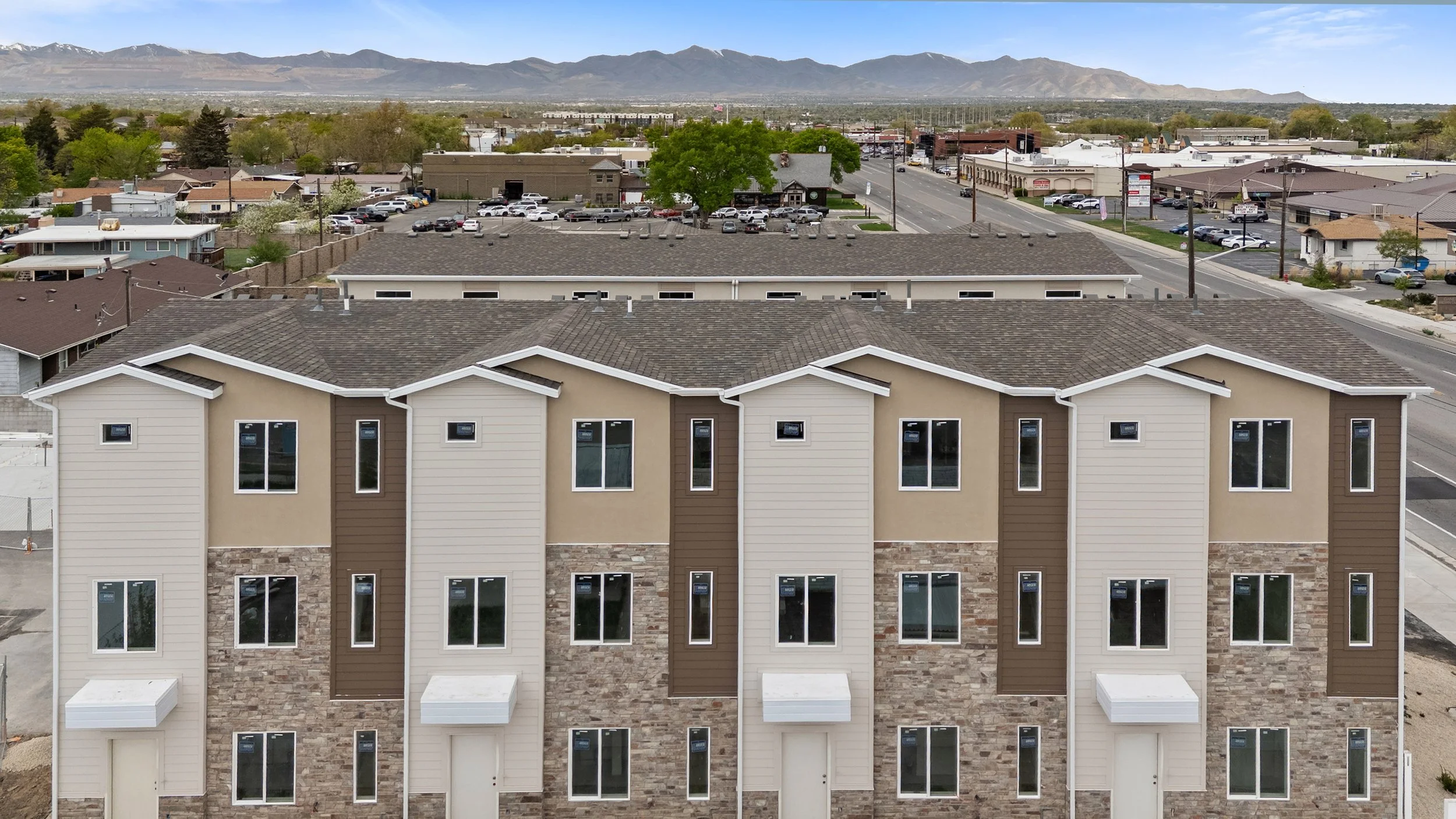 Front view of a multi-story residential building with beige siding and brick accents in an urban area. Multiple windows and garage doors are visible. In the background, there are commercial buildings, parking lots, and a mountain range under a partly cloudy sky.