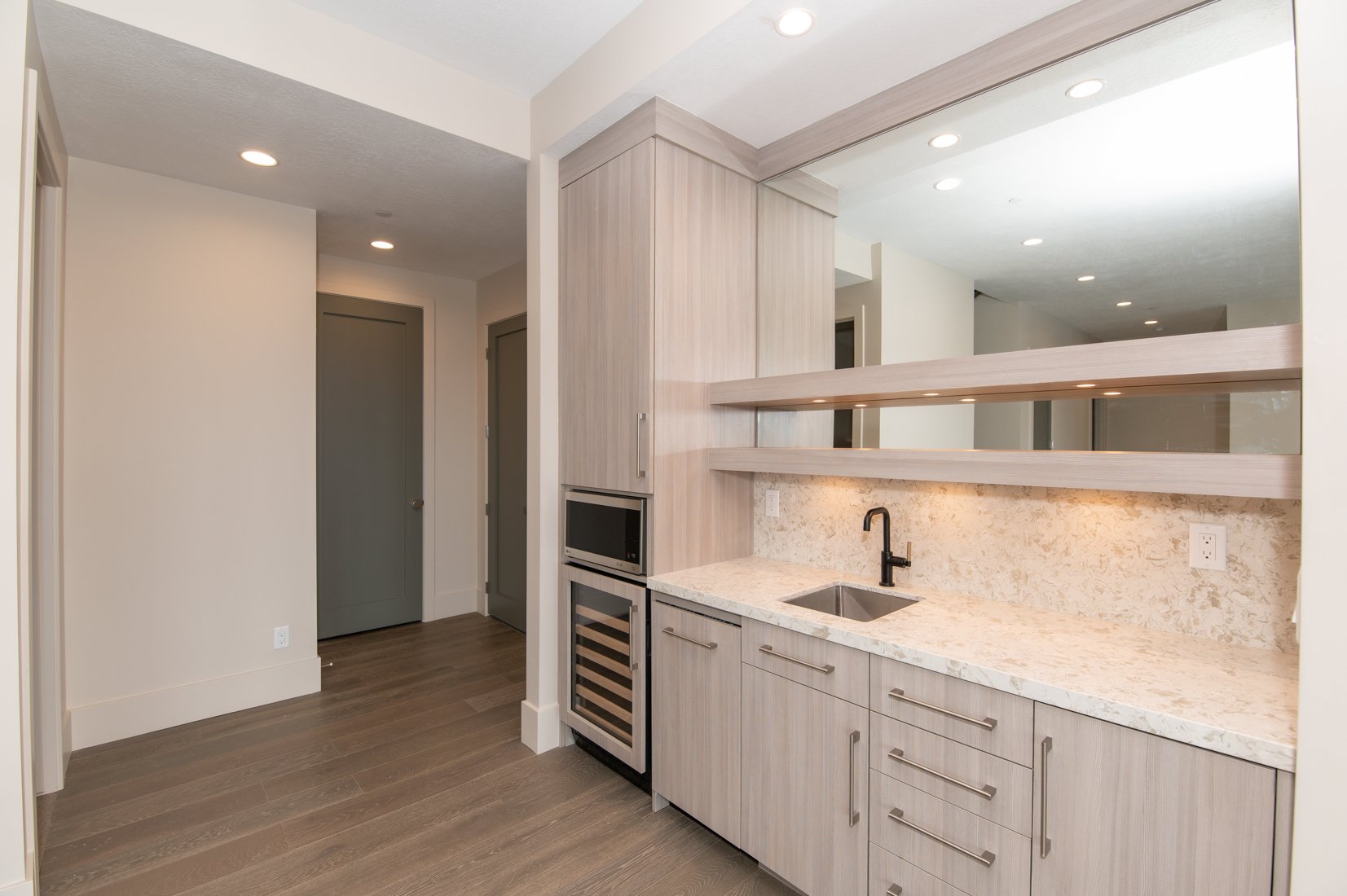 Modern kitchen with light wood cabinets, marble countertop, black faucet, built-in microwave and wine fridge, open shelves, and wood flooring.