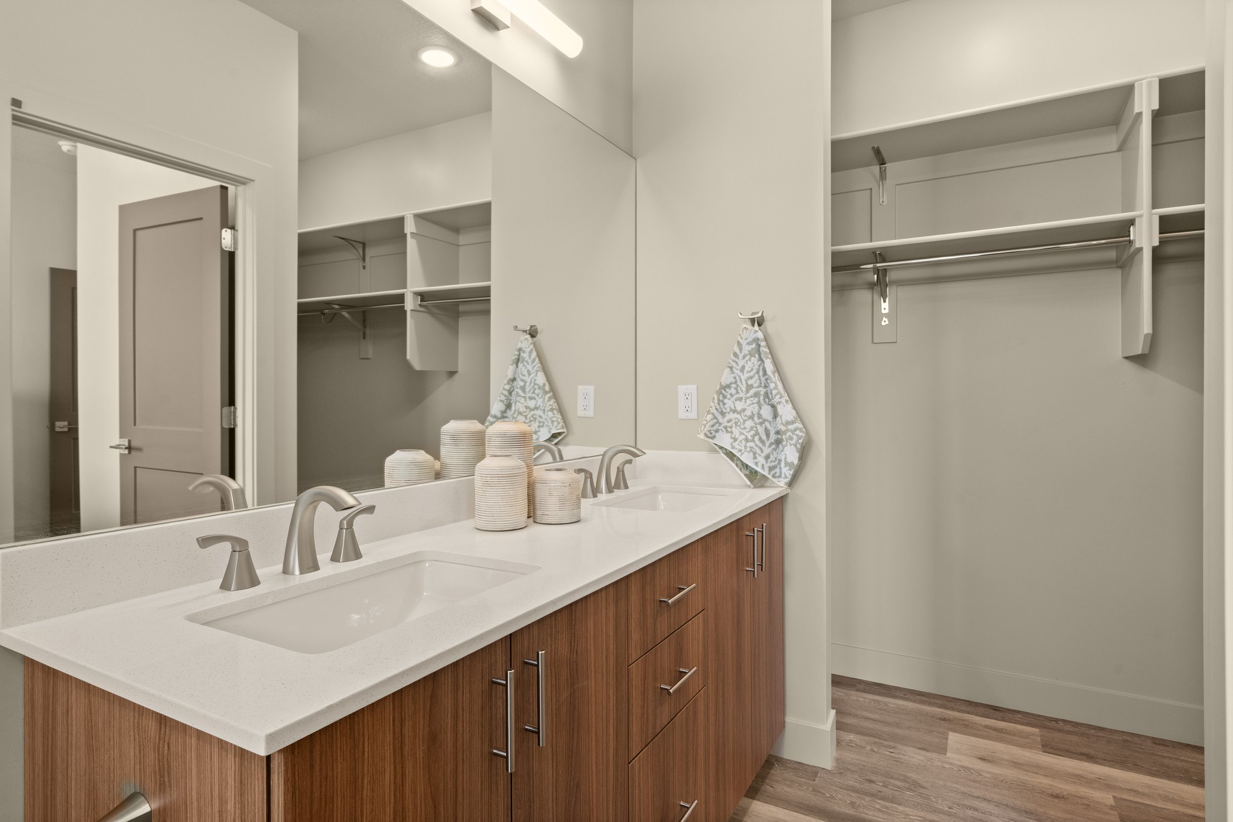 A modern bathroom vanity with a white countertop, dual sinks, and wooden cabinets. Above the sinks is a large mirror, and a few decorative towels and pottery are placed on the counter. There are open shelves and hanging rods for storage in the backgr