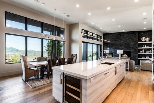 Modern open-concept kitchen and dining area with large windows, a white kitchen island, dining table with chairs, and a view of greenery outside.
