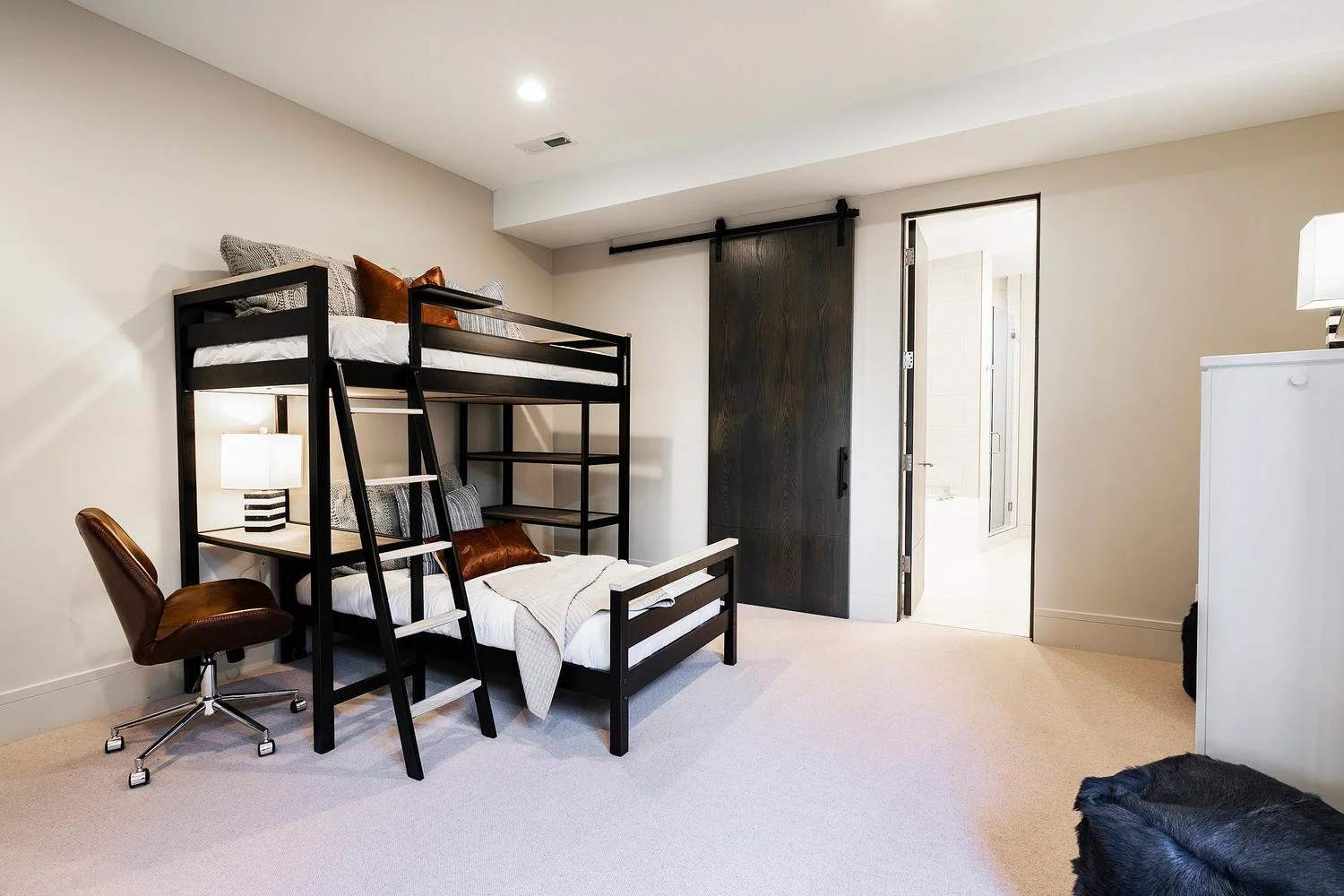 A bedroom with a bunk bed, desk chair, nightstand with lamp, and a dresser. The room has beige carpet and white walls, with a door leading to a bathroom.