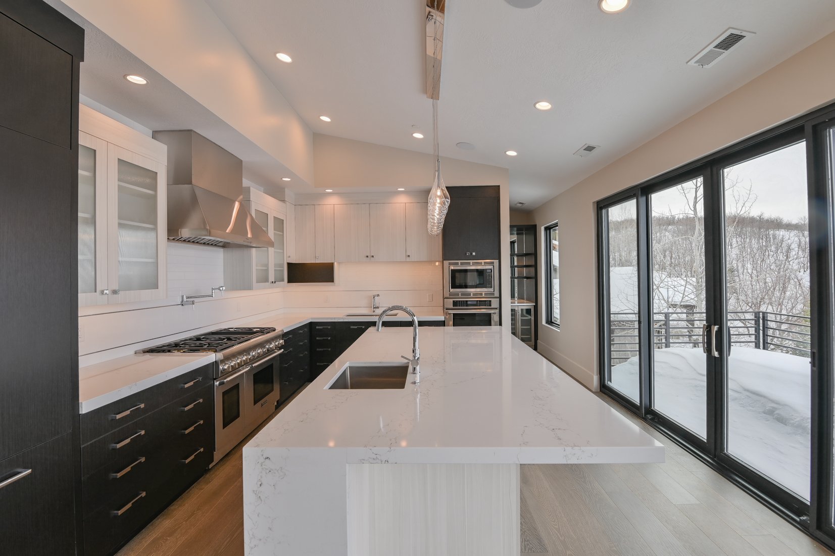 Modern kitchen with white countertops, black lower cabinets, white upper cabinets, stainless steel appliances, and large sliding glass doors leading to a snowy deck outside.