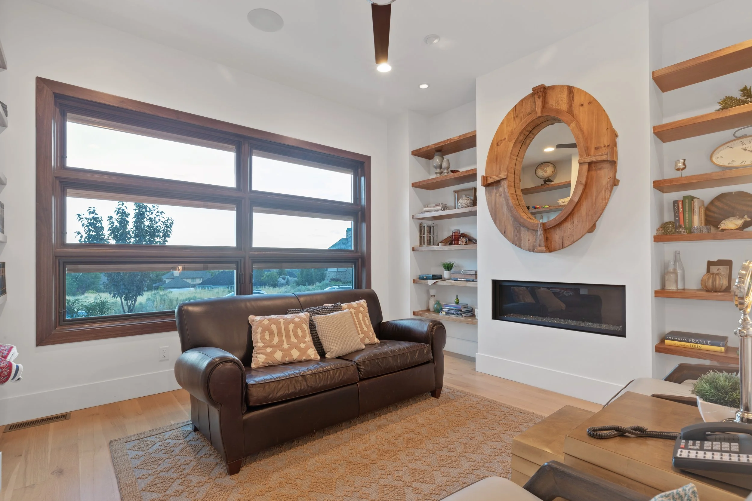 Living room with large window, leather sofa with decorative pillows, built-in shelves with books and decorative items, a round wooden mirror over a fireplace, and a wooden coffee table with a phone and a decorative object.