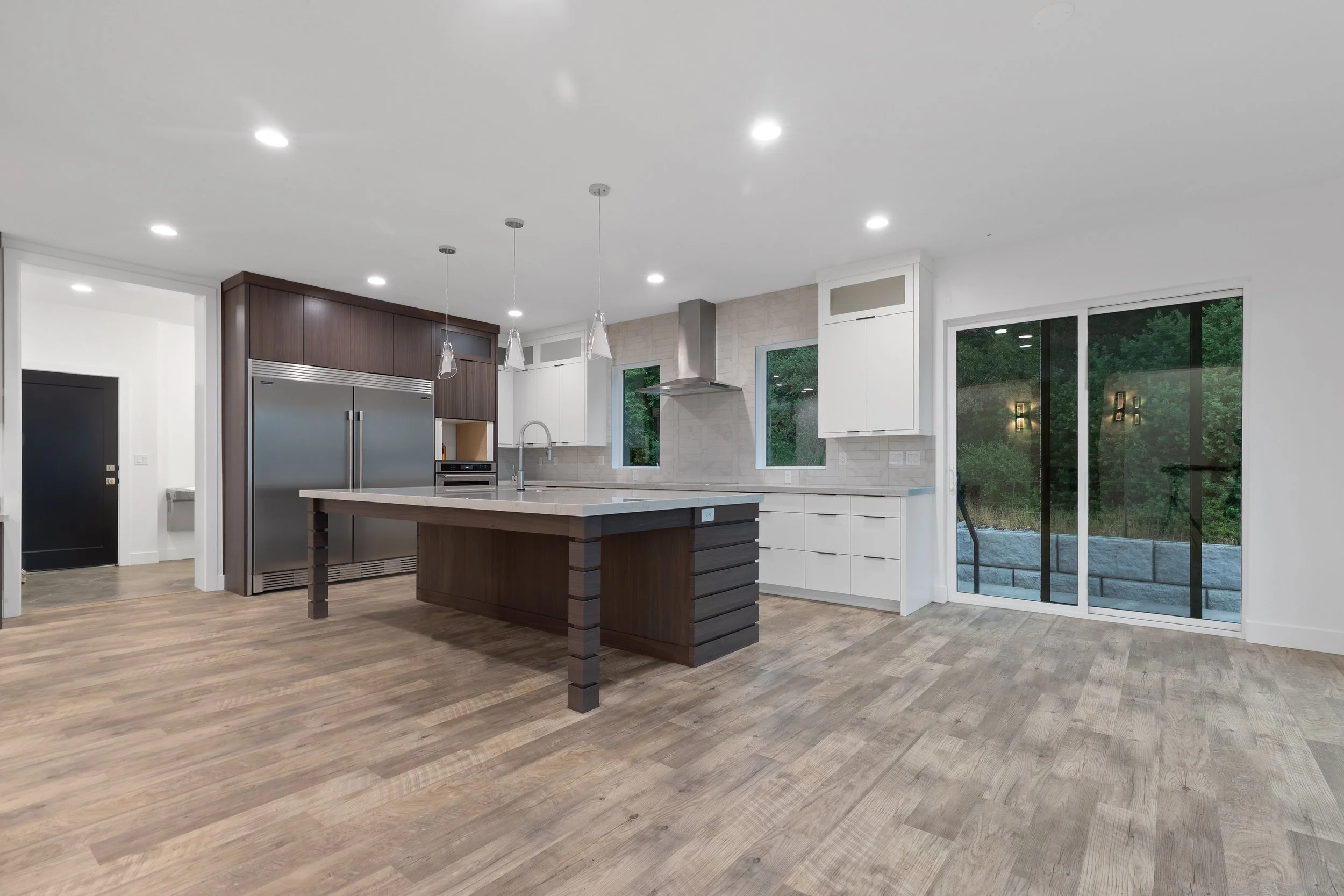 Modern kitchen with wooden flooring, white and dark brown cabinets, large stainless steel refrigerator, kitchen island with dark wood base and light countertop, pendant lights, and large sliding glass door overlooking a backyard.