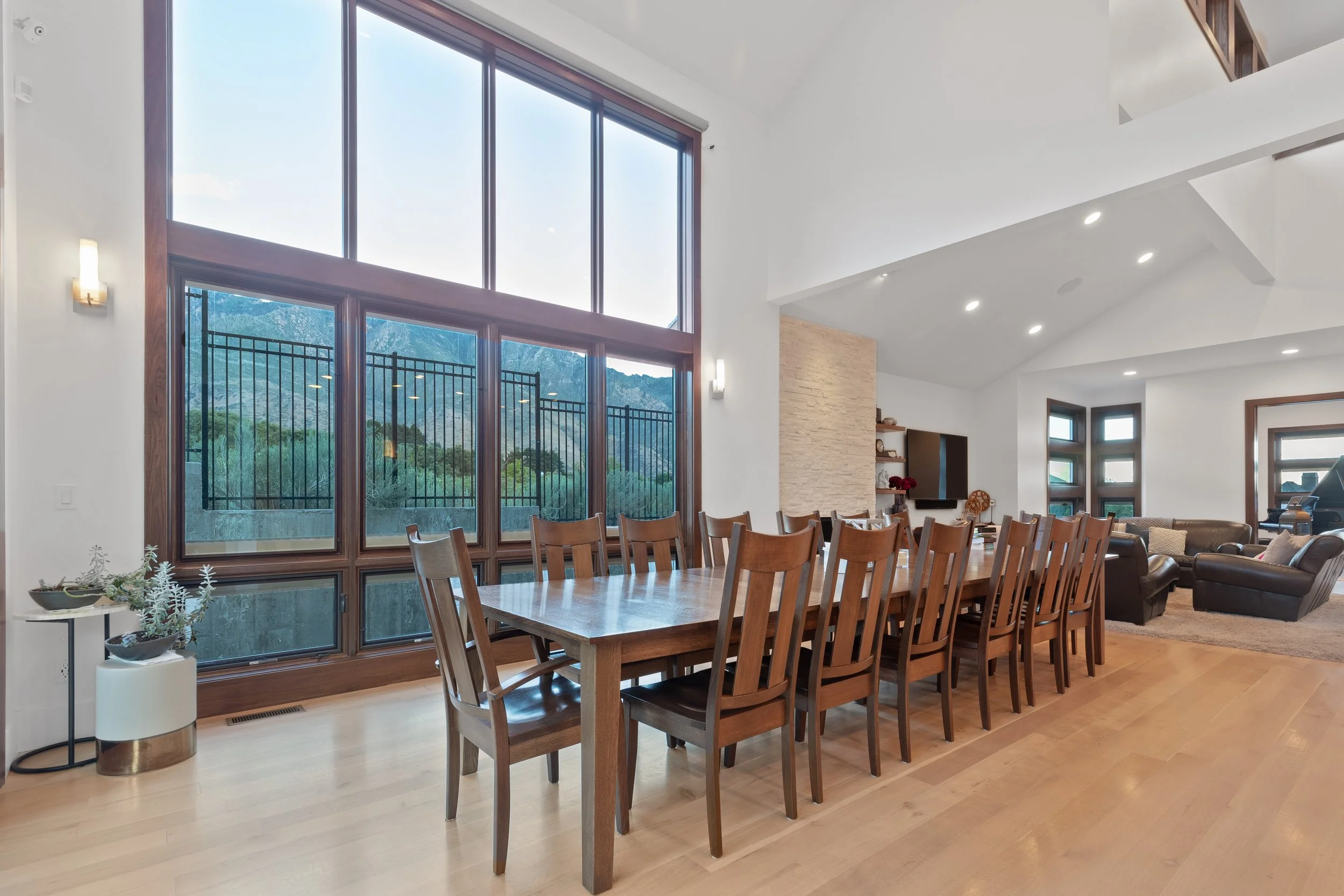 Spacious living area with large floor-to-ceiling windows showing a mountain landscape, a dining table with wooden chairs, a sitting area with sofas, a mounted TV, and modern decor.