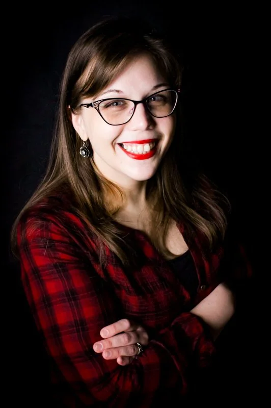 A young woman with shoulder-length brown hair, glasses, and red lipstick, smiling and crossing her arms, wearing a red plaid shirt against a dark background.