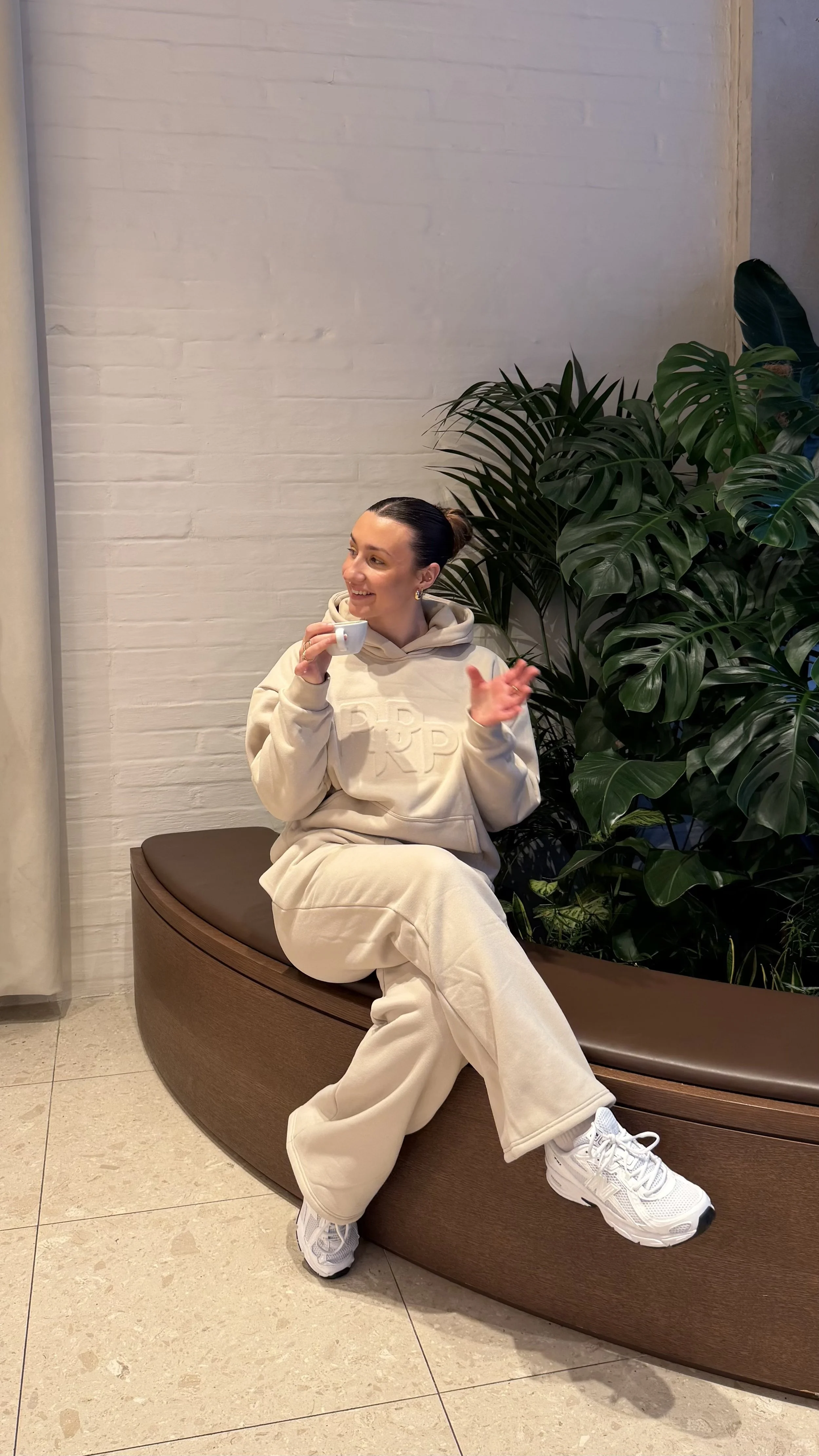 A woman sitting on a rounded wooden bench, holding a small cup, smiling and looking to the left, with large green plants behind her, in a modern indoor setting with a white brick wall.
