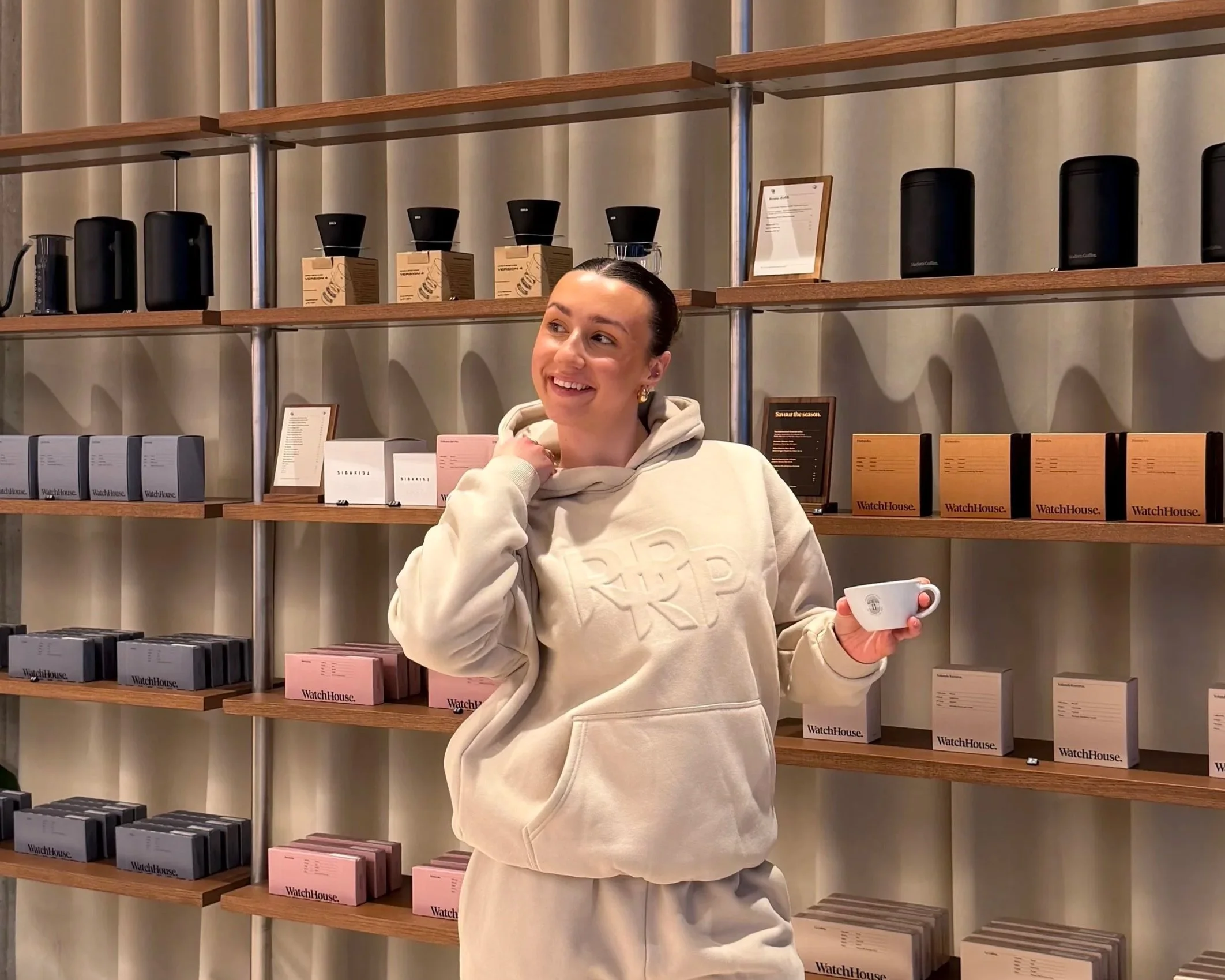 A woman smiling while shopping in a store, holding a white mug with a store brand logo, standing in front of wooden shelves with boxes of watches and coffee equipment.