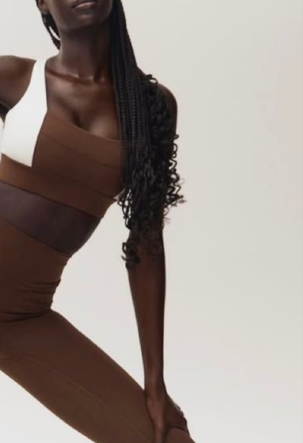 Close-up of a woman with long curled hair, wearing a color-blocked gym set in white, brown, and black, against a plain background.