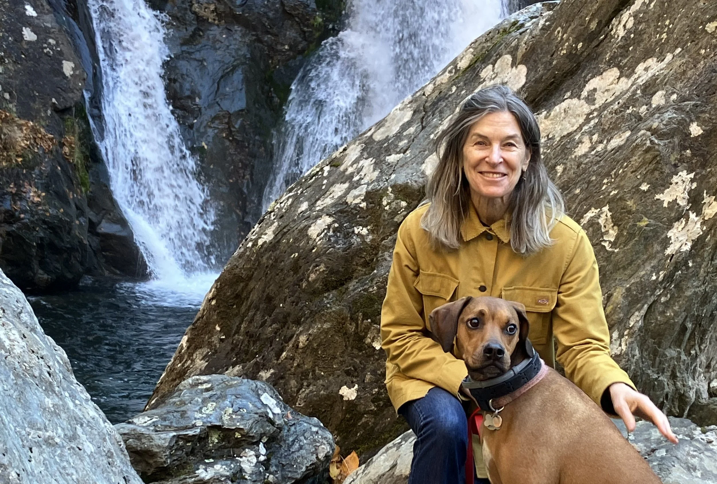 A smiling woman with gray hair wearing a yellow jacket, sitting on rocks next to a dog with a waterfall in the background.