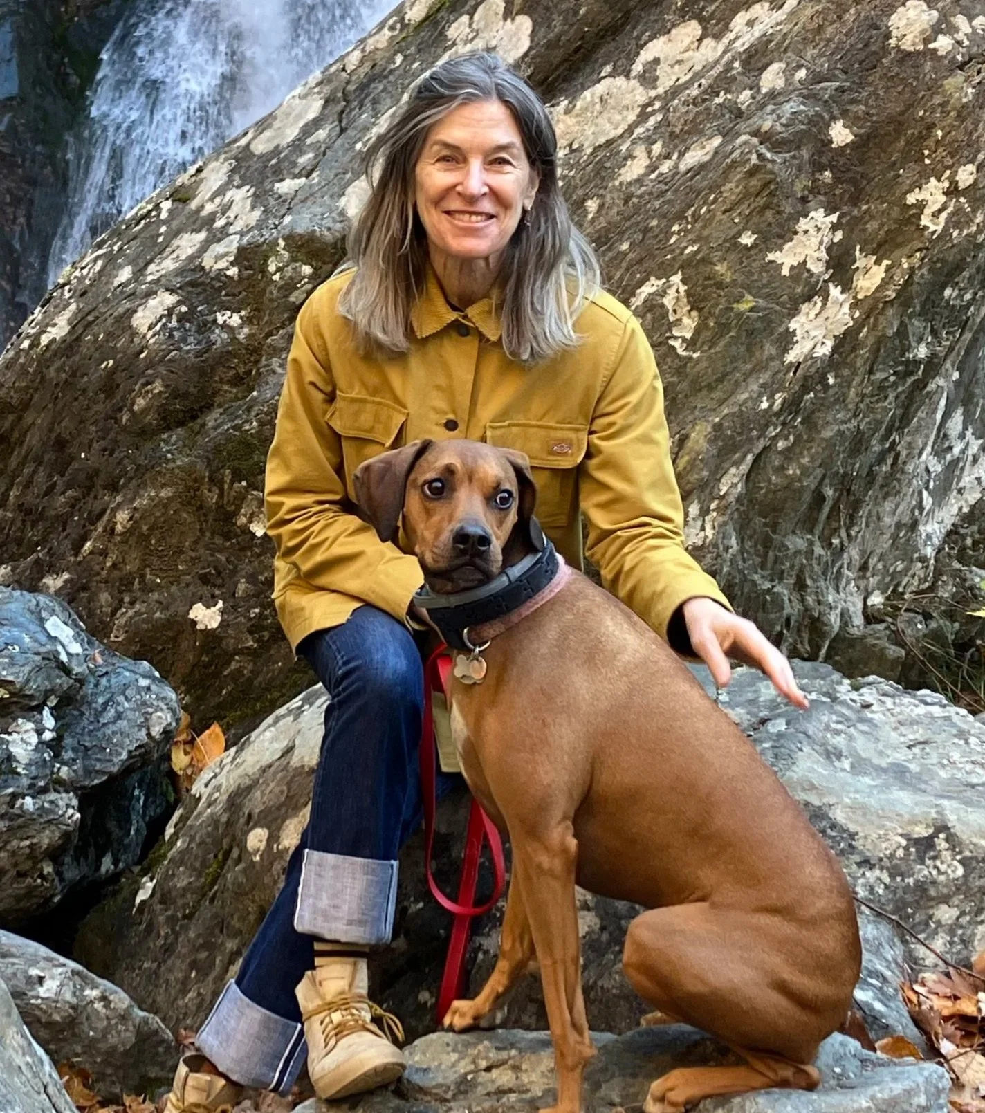 A woman in a yellow jacket and jeans sitting on rocks with her brown dog, near a waterfall in a rocky outdoor setting.