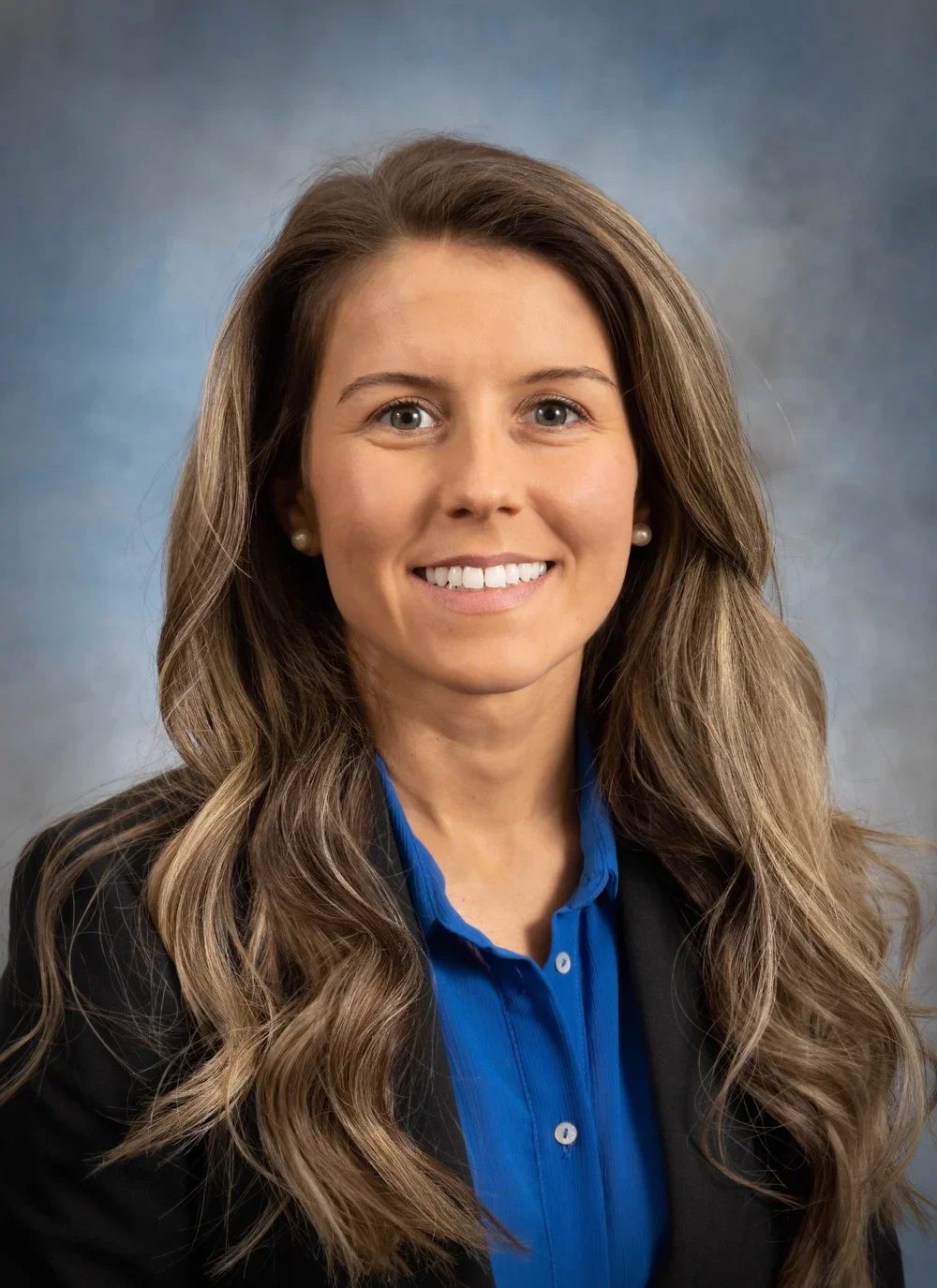 A professional woman with long wavy brown hair wearing a black blazer with a blue blouse, smiling against a neutral blue-grey background.