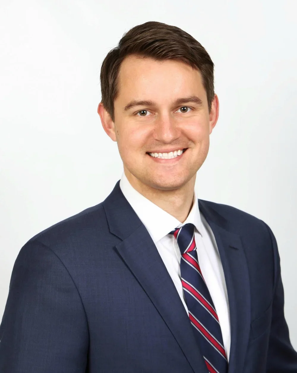 A young man in a navy suit with a white shirt and a striped red, white, and navy tie, smiling against a plain white background.