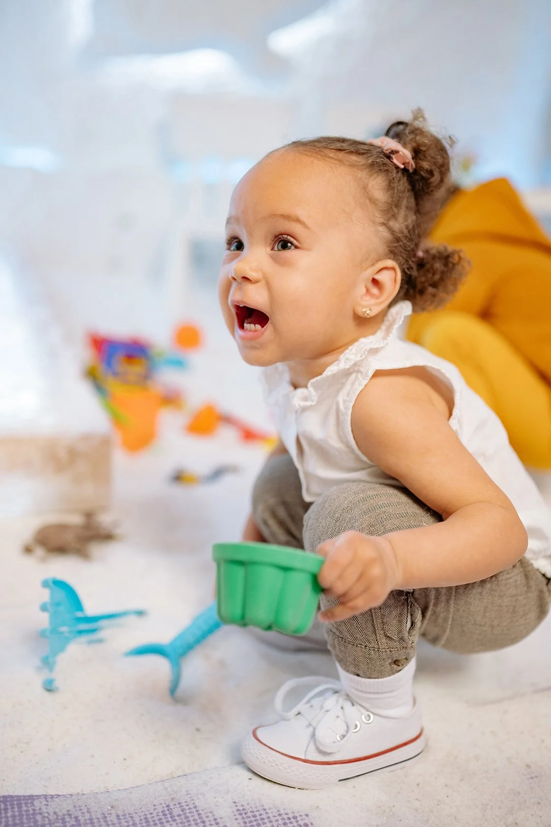 A young girl with curly hair in a ponytail, squatting on a play mat inside a bright room. She is playing with colorful toys during in-home speech therapy for kids in Redondo Beach, CA