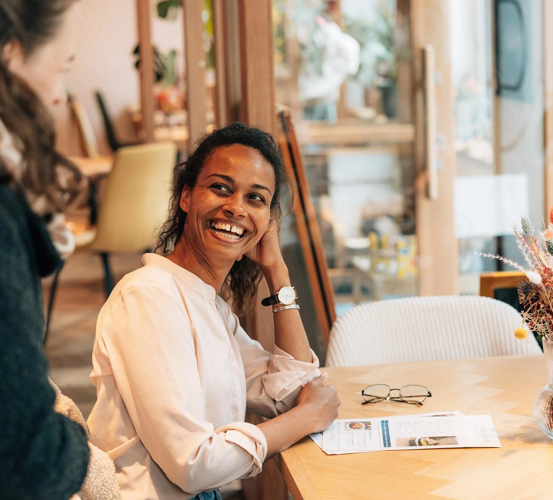 Zwei Frauen im Gespräch an einem Cafétisch, eine von ihnen lacht herzlich.