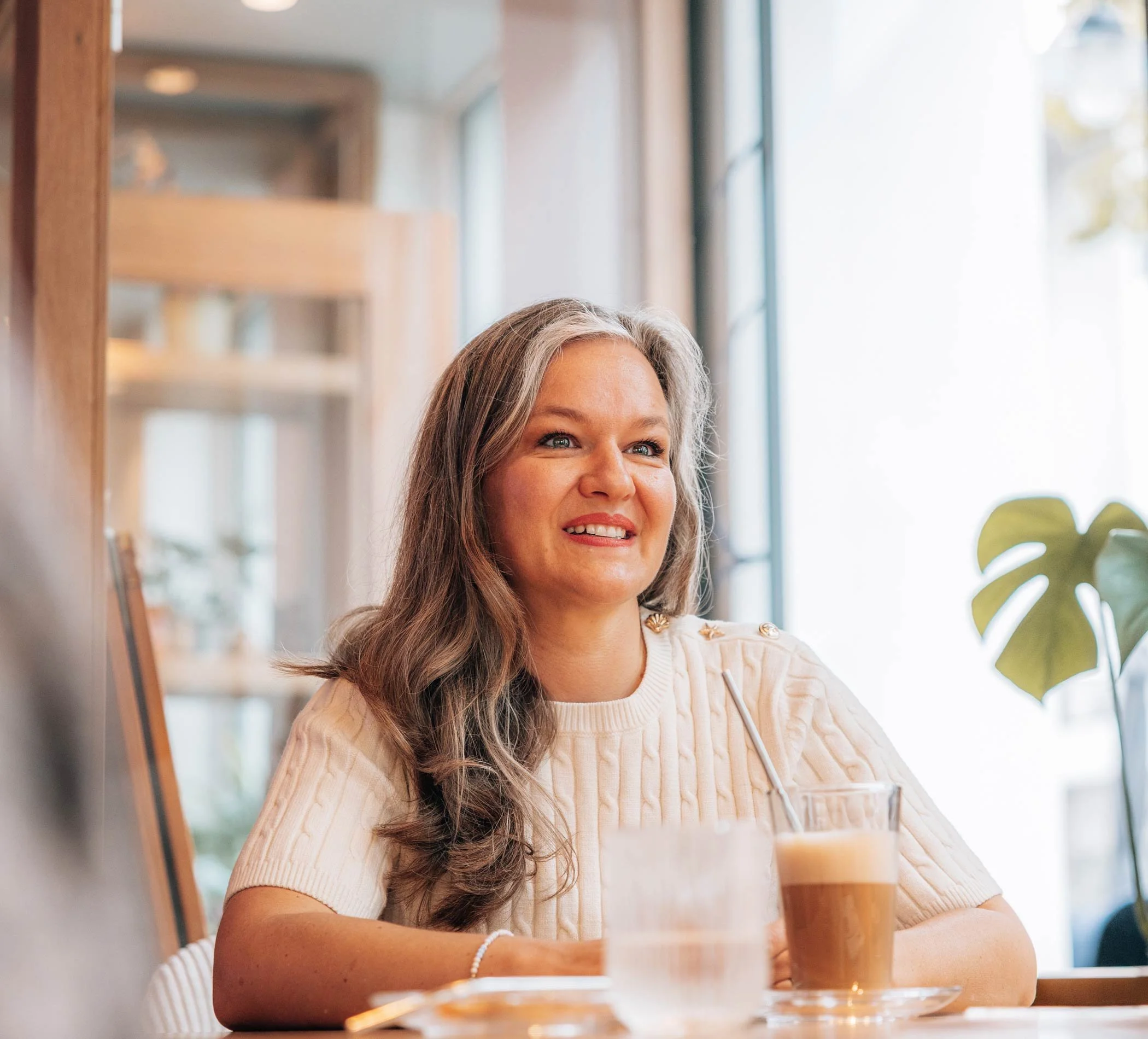 Eine Frau mit langen grauen Haaren sitzt lächelnd mit einem Milchkaffee im Café.