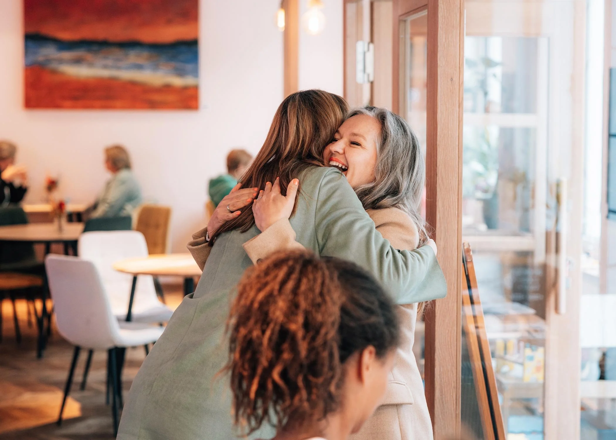 Zwei Frauen umarmen sich lachend in einem Café, im Vordergrund sitzt eine weitere Frau.
