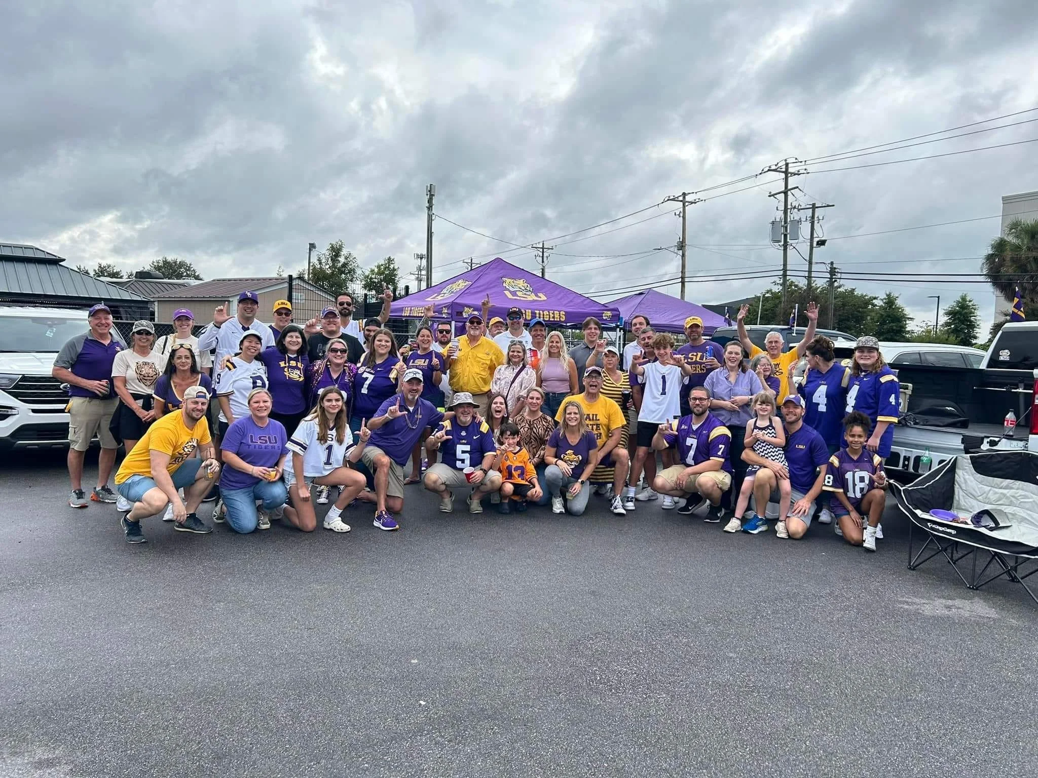 A large group of LSUY fans gathered at the LSU v SC 2024 game in Columbia SC, wearing purple and gold clothing and LSU Tigers gear, in front of a purple LSU Tigers tent, celebrating and posing for a group photo on a cloudy day.