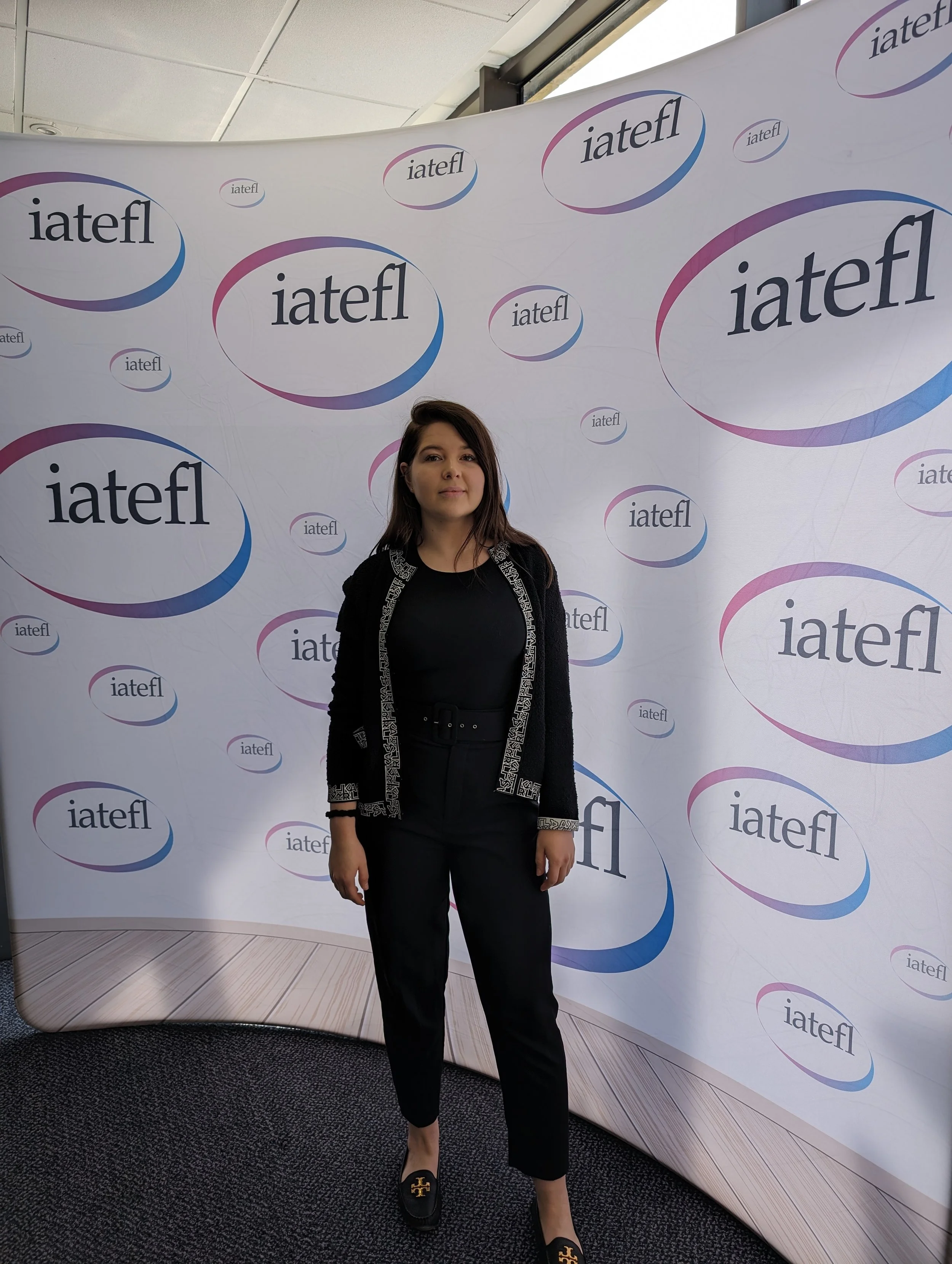 Elizabeth Demine standing at an IATEFL conference in a black outfit, in front of a backdrop featuring the IATEFL logo.
