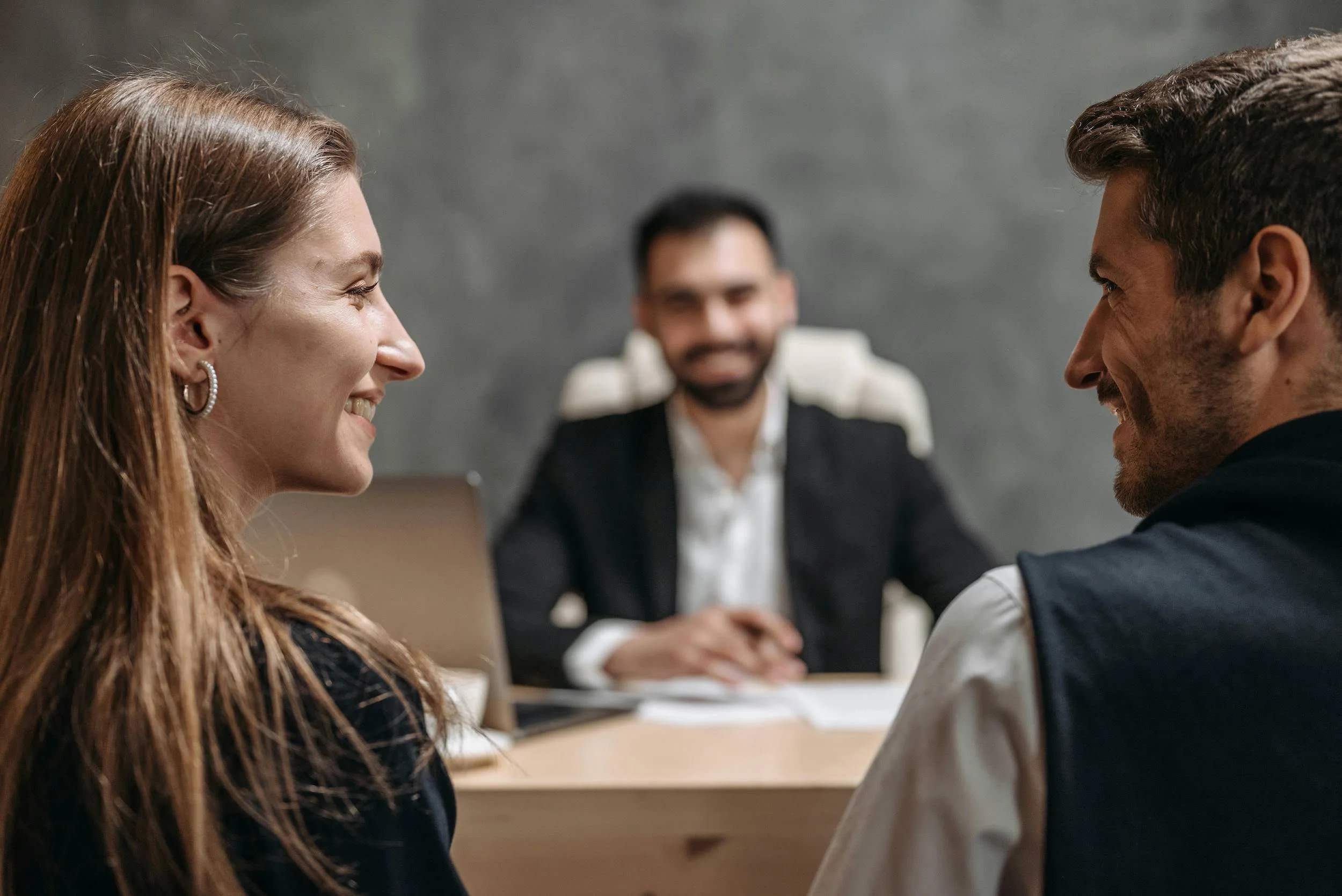 A woman and a man are facing each other and smiling in close proximity, likely in an office or meeting room. A third person, a man, is visible in the background, slightly out of focus, sitting at a table.