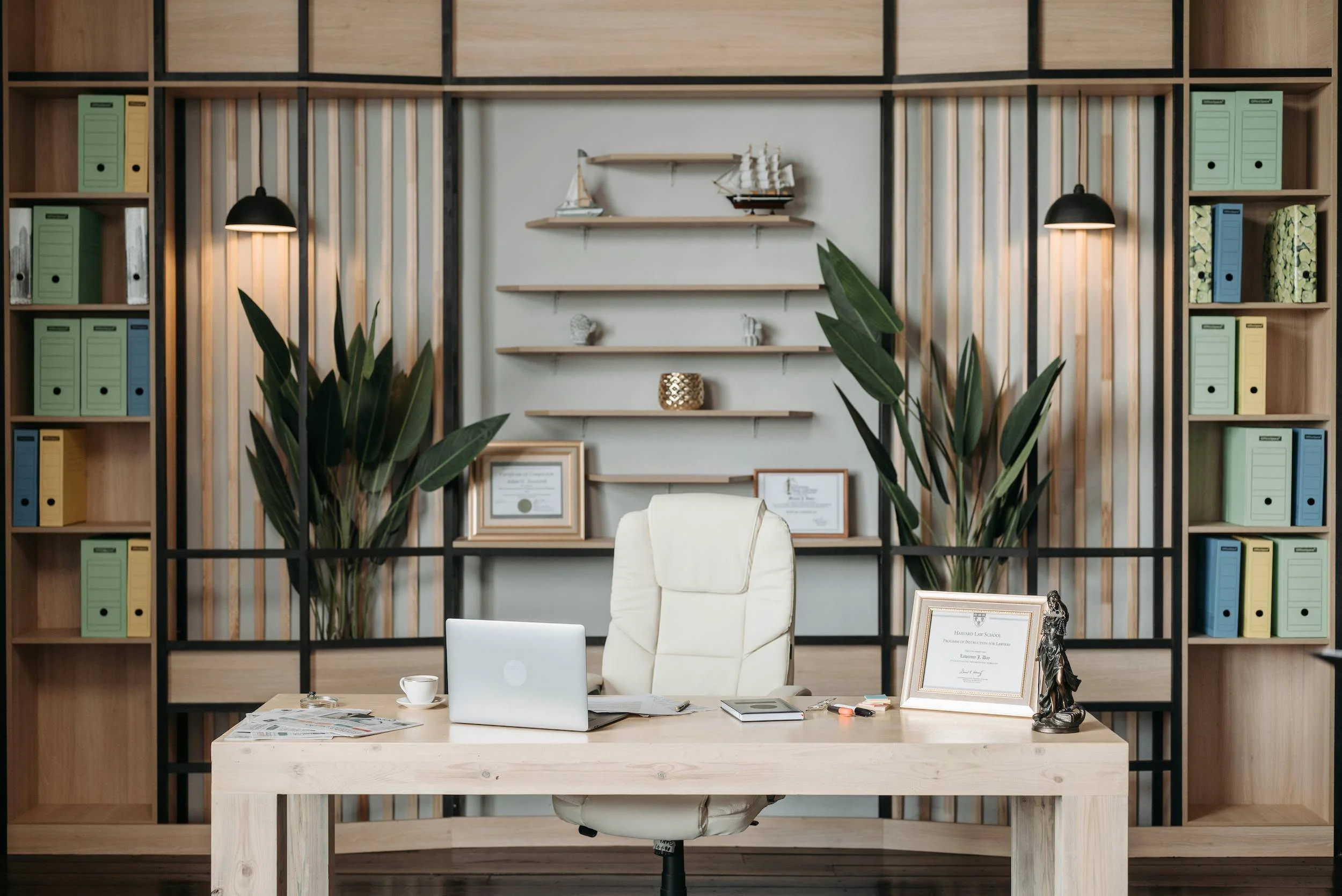 A workspace with a wooden desk, an office chair, a laptop, newspapers, a cup, and framed documents. Behind the desk is a bookshelf with colored binders, decorative items, and potted plants, with shelves on the wall holding model ships and small sculptures.