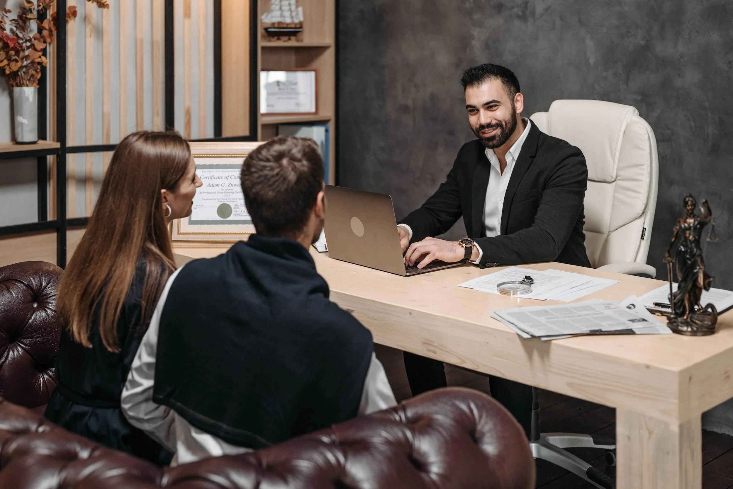 A man in a black suit and white shirt sitting at a desk, smiling, with two people in front of him, a woman with long red hair and a man with short brown hair, in an office with dark walls, wooden shelves, and framed certificates.
