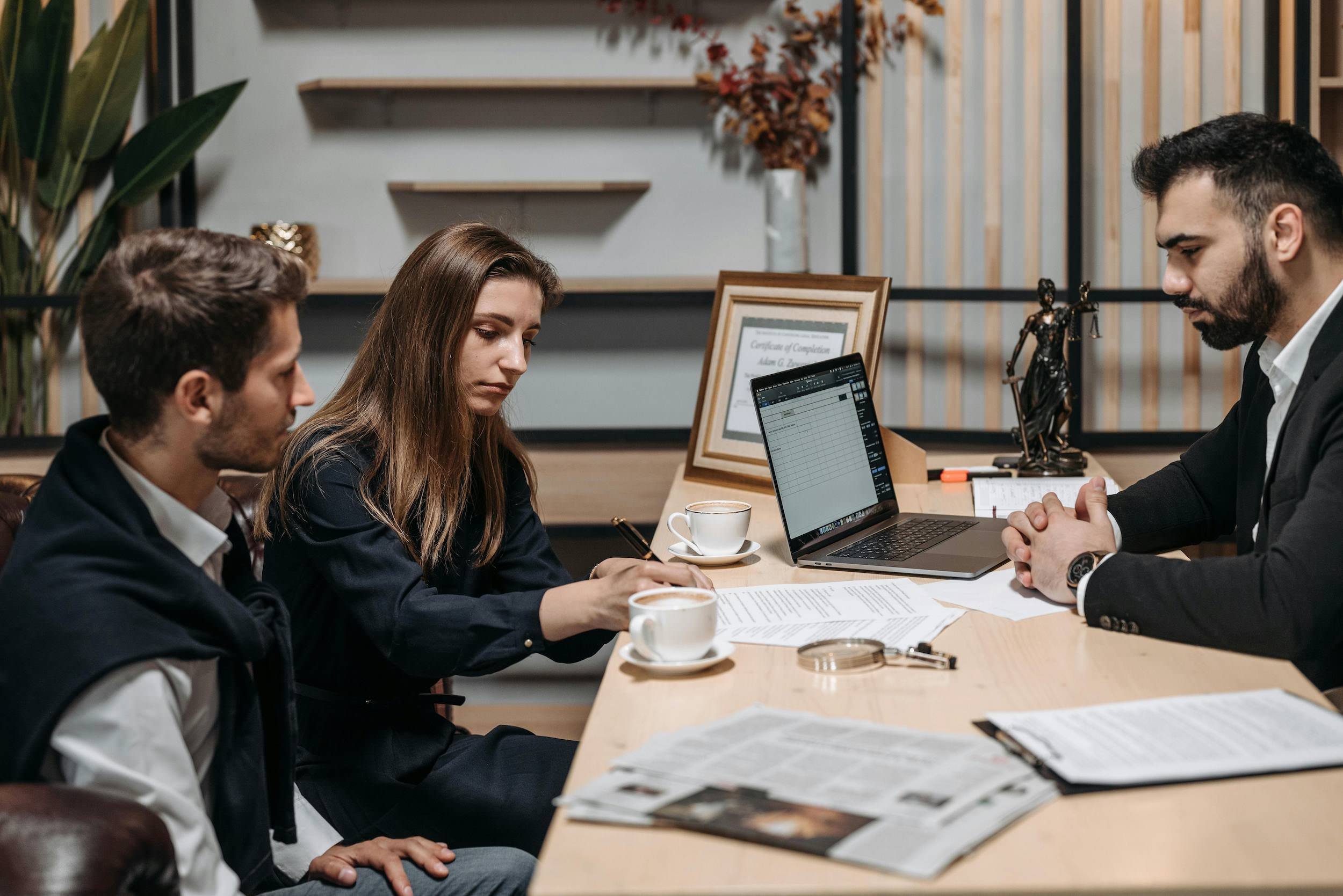 Four people sitting at a conference table engaged in a discussion. Papers, notebooks, coffee cups, and a laptop are on the table. One man on the right is looking at the laptop, while a woman is taking notes. The setting appears to be an office or meeting room with decorative items and wooden shelves in the background.