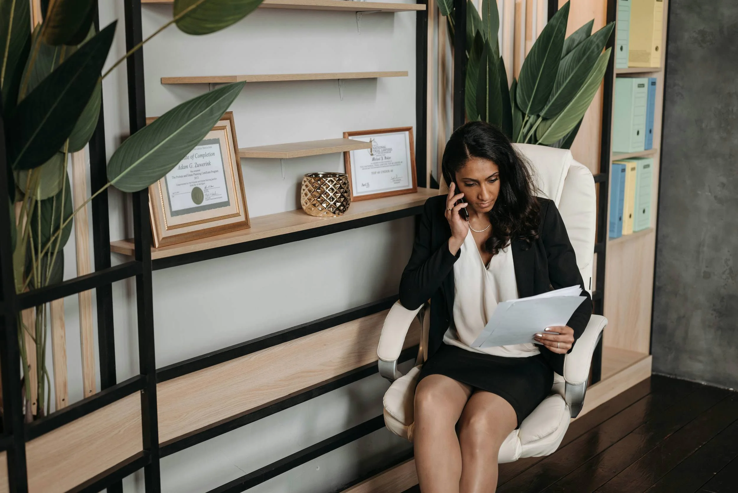 Businesswoman sitting in a white office chair, talking on her mobile phone, holding a document, with framed certificates on a shelf behind her, and large green plants in the background.