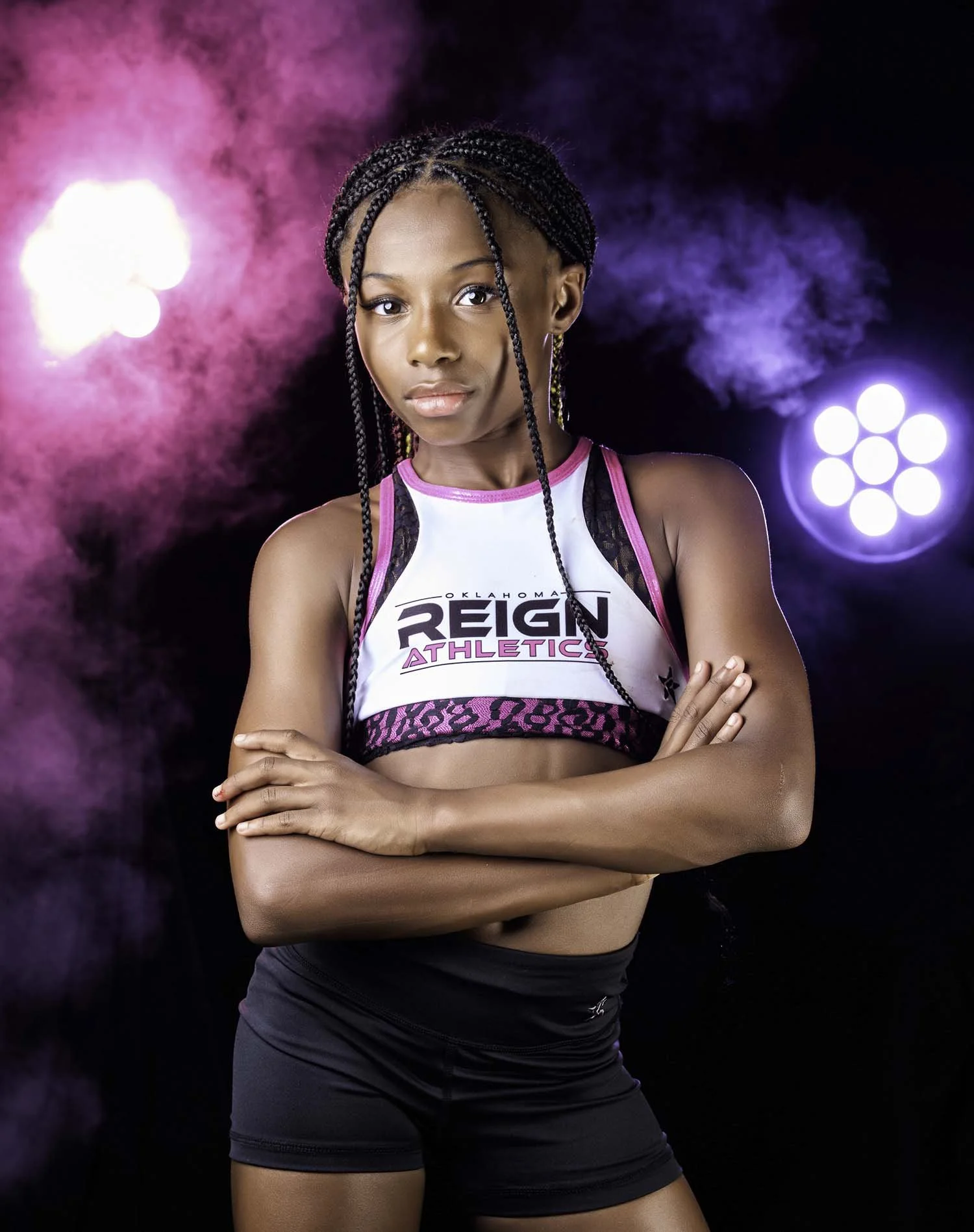 A young female athlete with braided hair, wearing a white and black athletic crop top with pink accents, standing confidently with crossed arms against a colorful, smoky background with pink and purple lights.