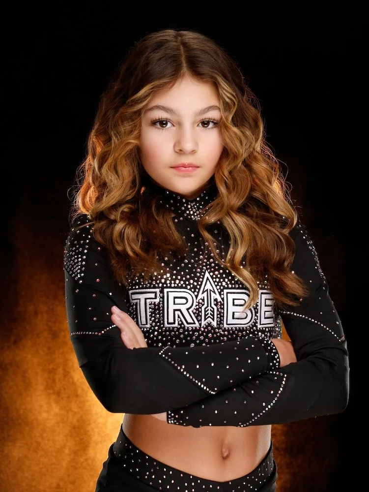 Young girl with long, wavy hair, wearing a black sports crop top with the word 'TRIBE' and an arrow design, covered in rhinestones, standing with arms crossed against a black and orange gradient background.