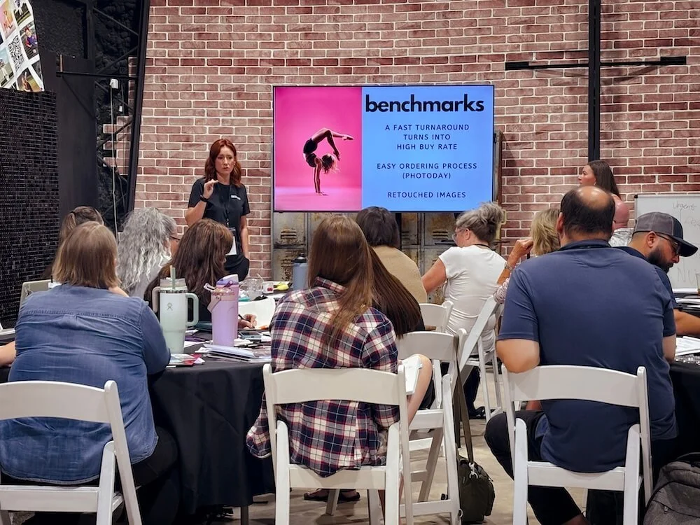 A woman giving a presentation to a group of people seated at tables in a room with brick walls. A screen behind her displays the word 'benchmarks' and some presentation points.