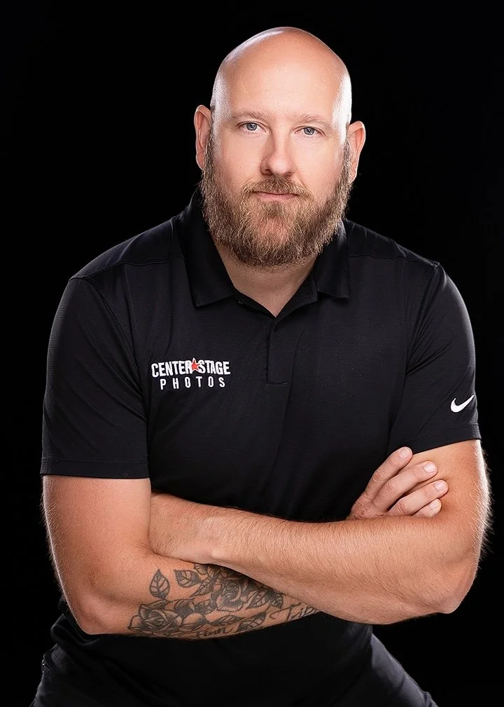 Portrait of a bald man with a beard, wearing a black polo shirt with a logo that reads 'Center Stage Photos,' posing with arms crossed against a black background.