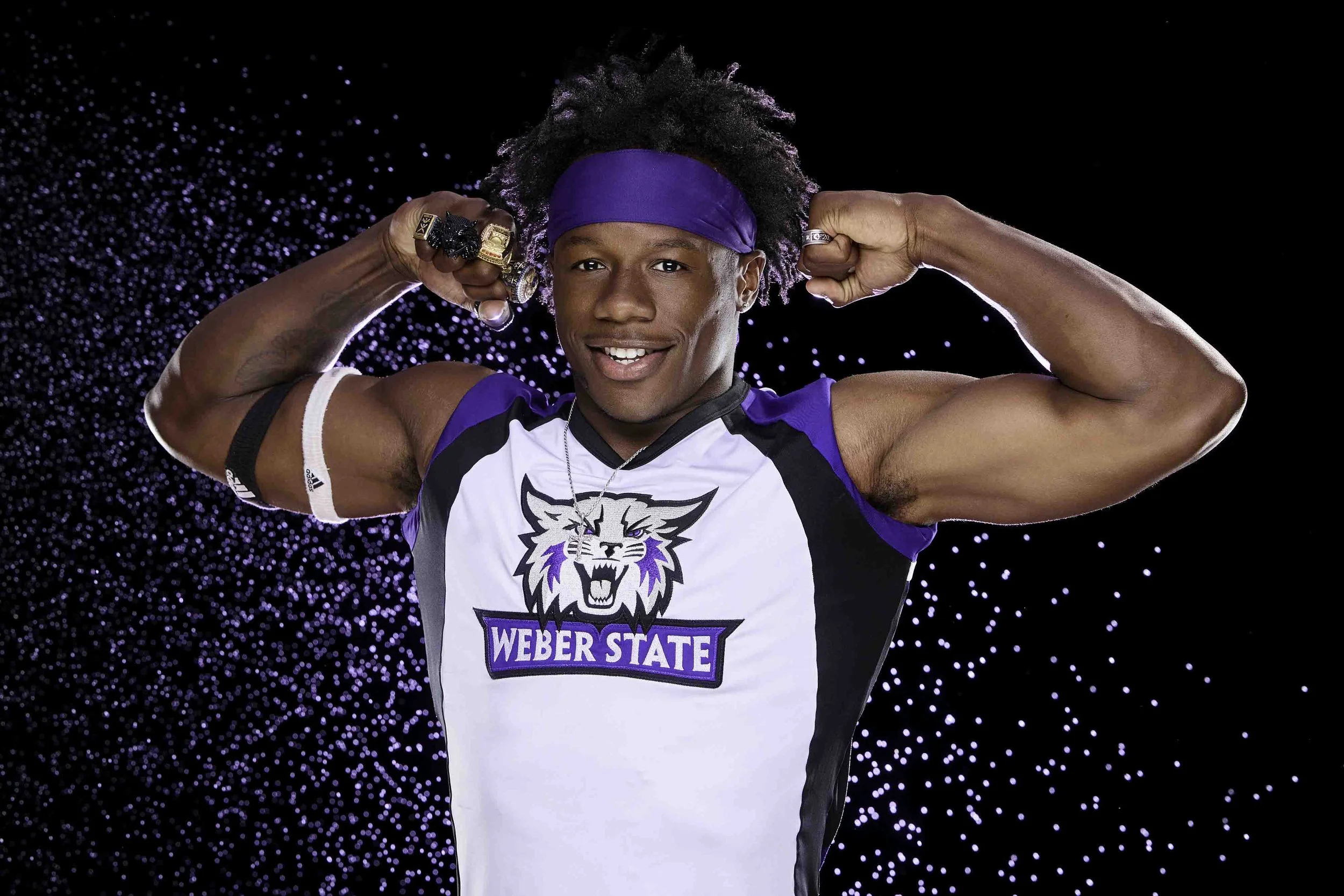 Young man with dressed hair, wearing a purple headband, smiling and flexing his biceps, wearing a sports jersey with the logo of Weber State Wildcats, standing in front of a black background with purple and white dots.