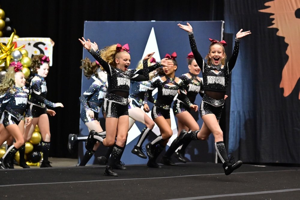 Cheerleaders in matching black, white, and pink uniforms with 'Top Gun' on the front, performing a dance routine on stage during a cheerleading competition, with some members jumping and smiling.