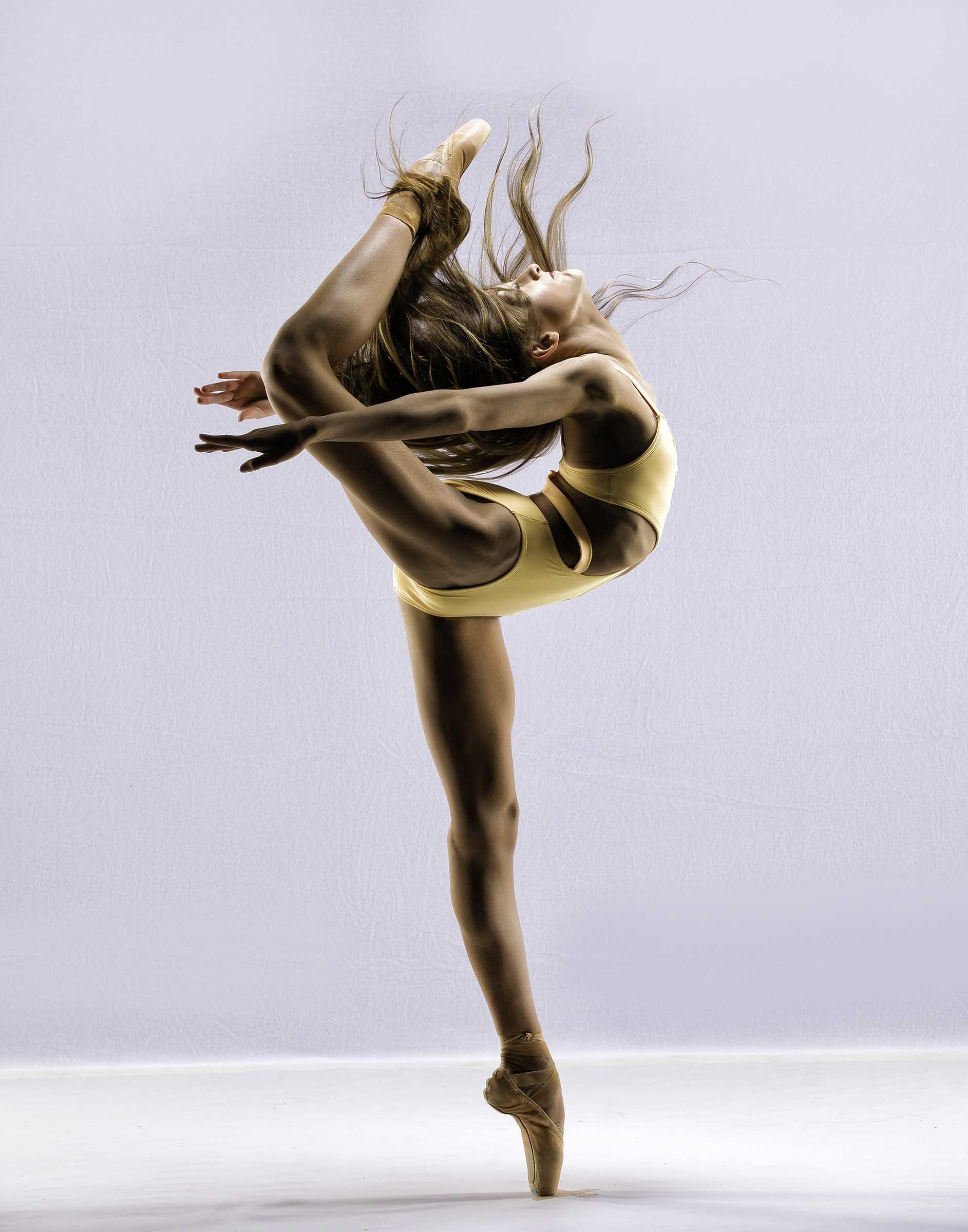 A ballet dancer is performing a standing split on pointe, with one leg extended upward and the other supporting her on pointe, against a plain light background.