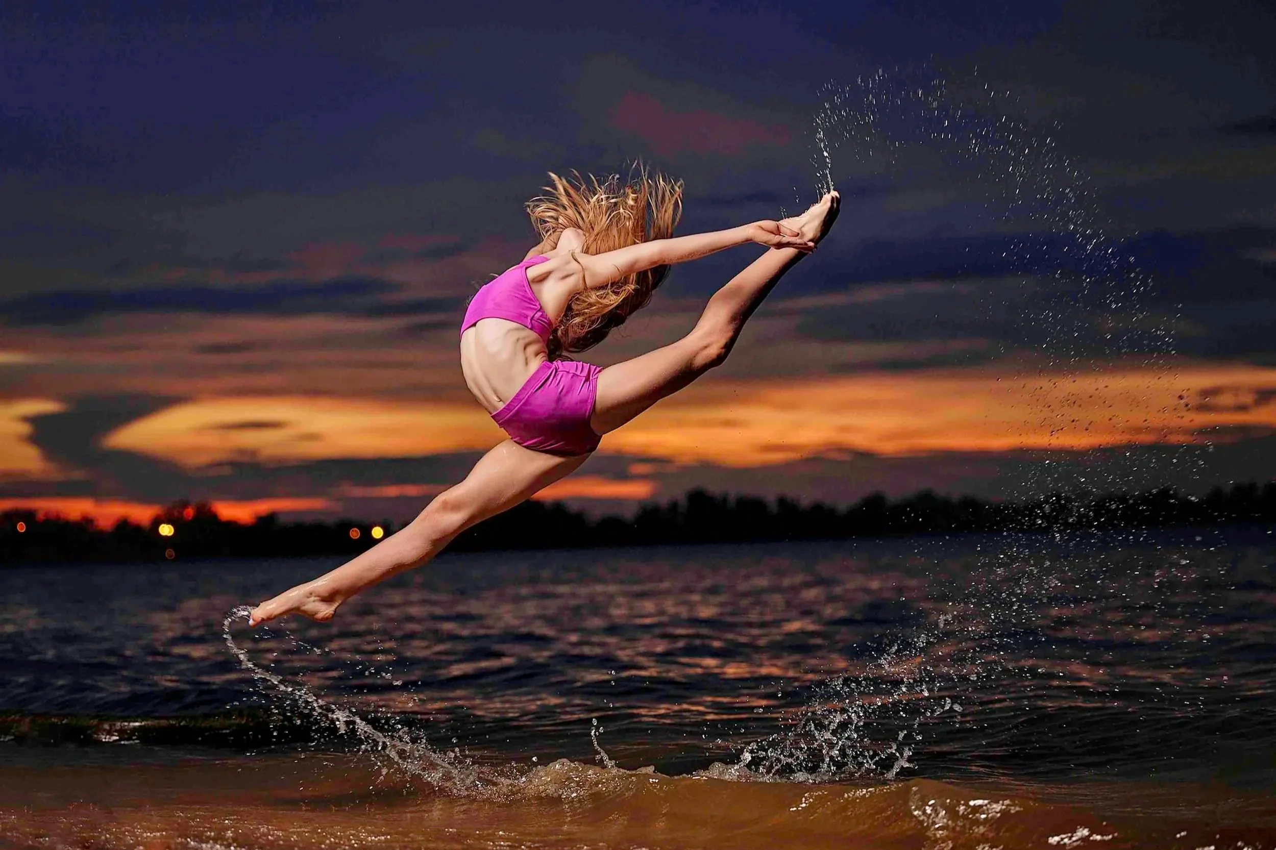 A woman in a pink outfit is jumping off a dock into a body of water at sunset, with her arms stretched forward and water splashing around her.