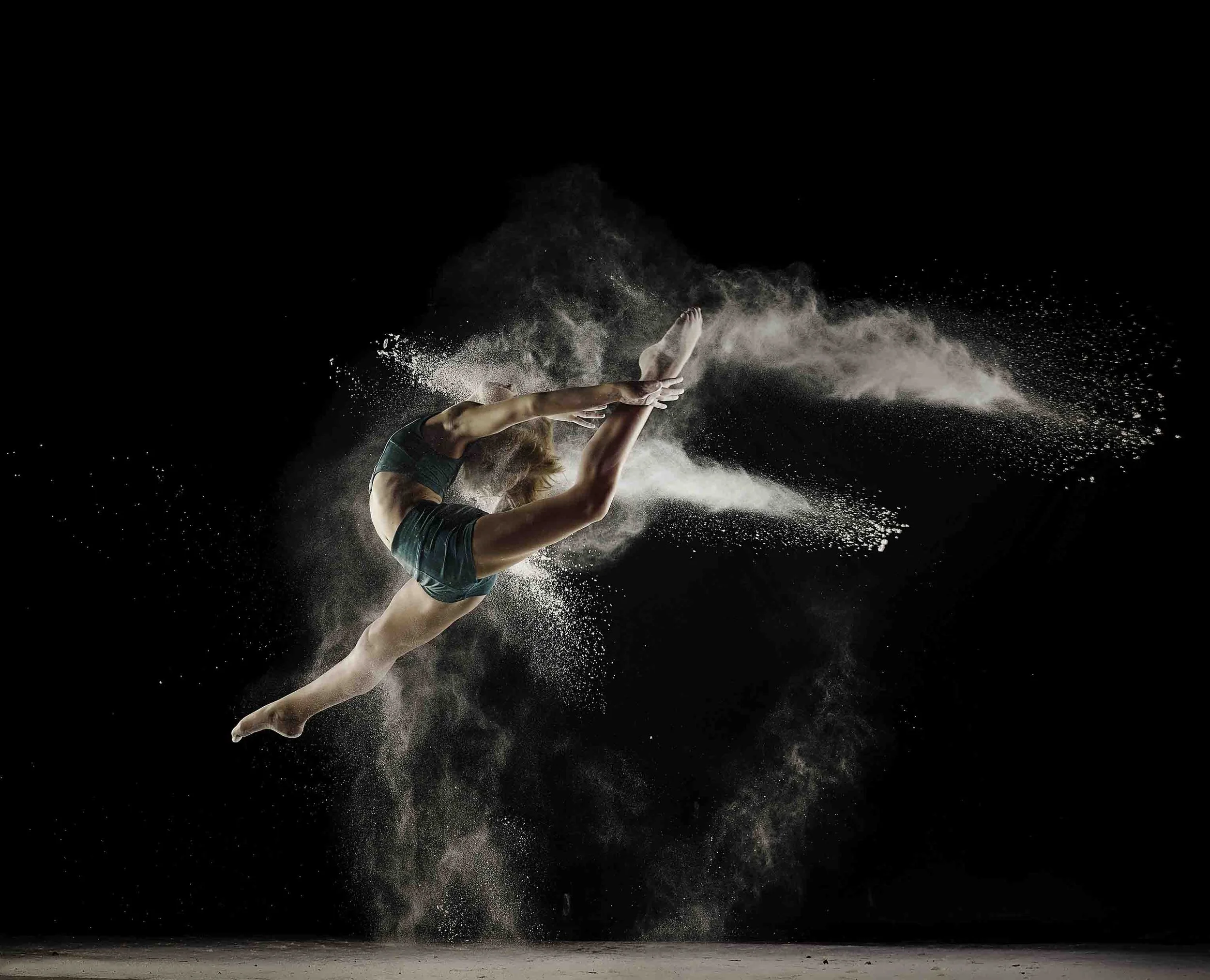A female gymnast performing a leap in the air with a splash of chalk or powder around her, against a black background.