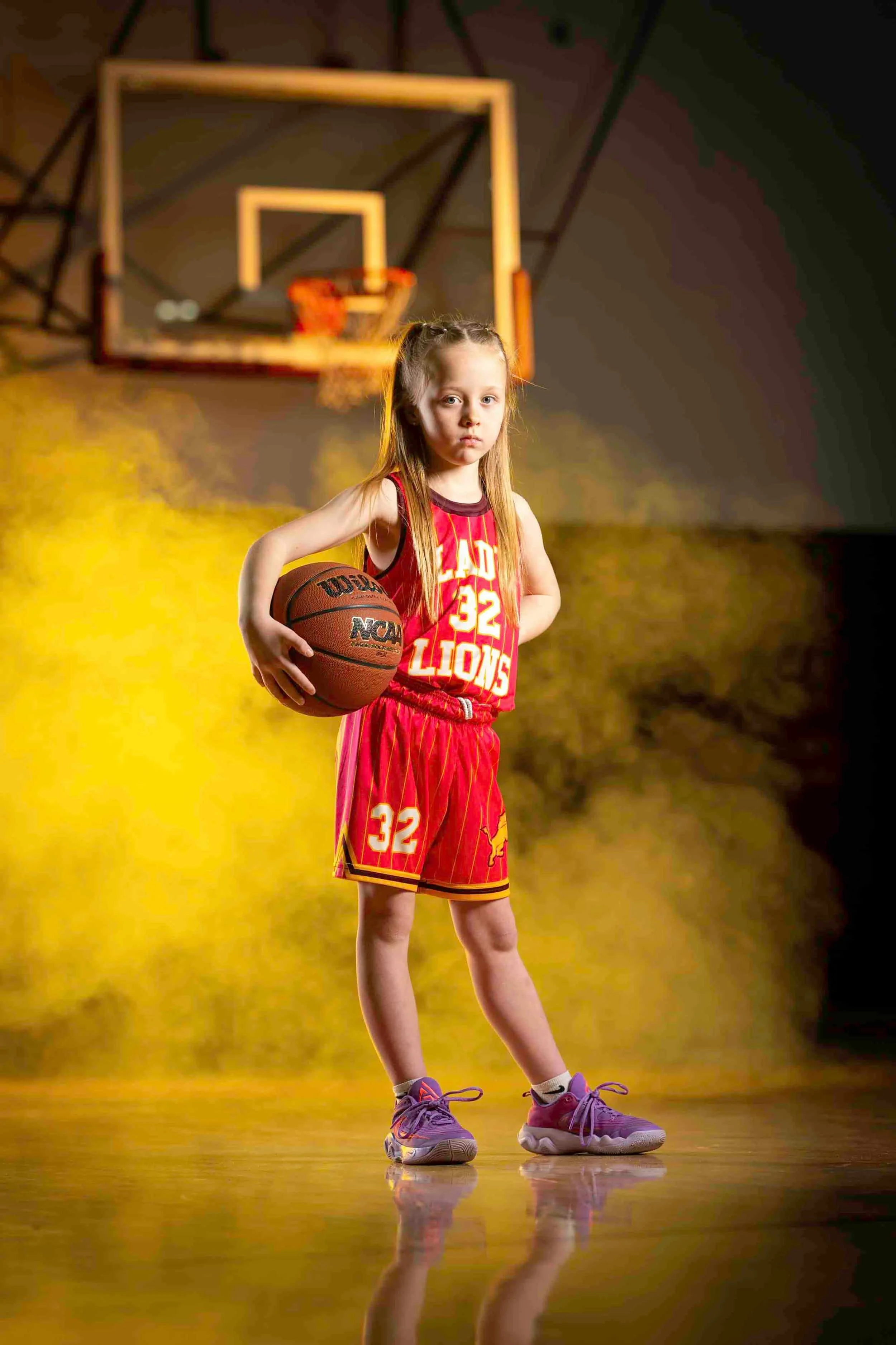 A young girl with long blonde hair in a ponytail, wearing a red basketball uniform with the number 32 and the word 'Lions', stands on a basketball court holding a basketball in her right hand. There is a basketball hoop and backboard in the backgroun